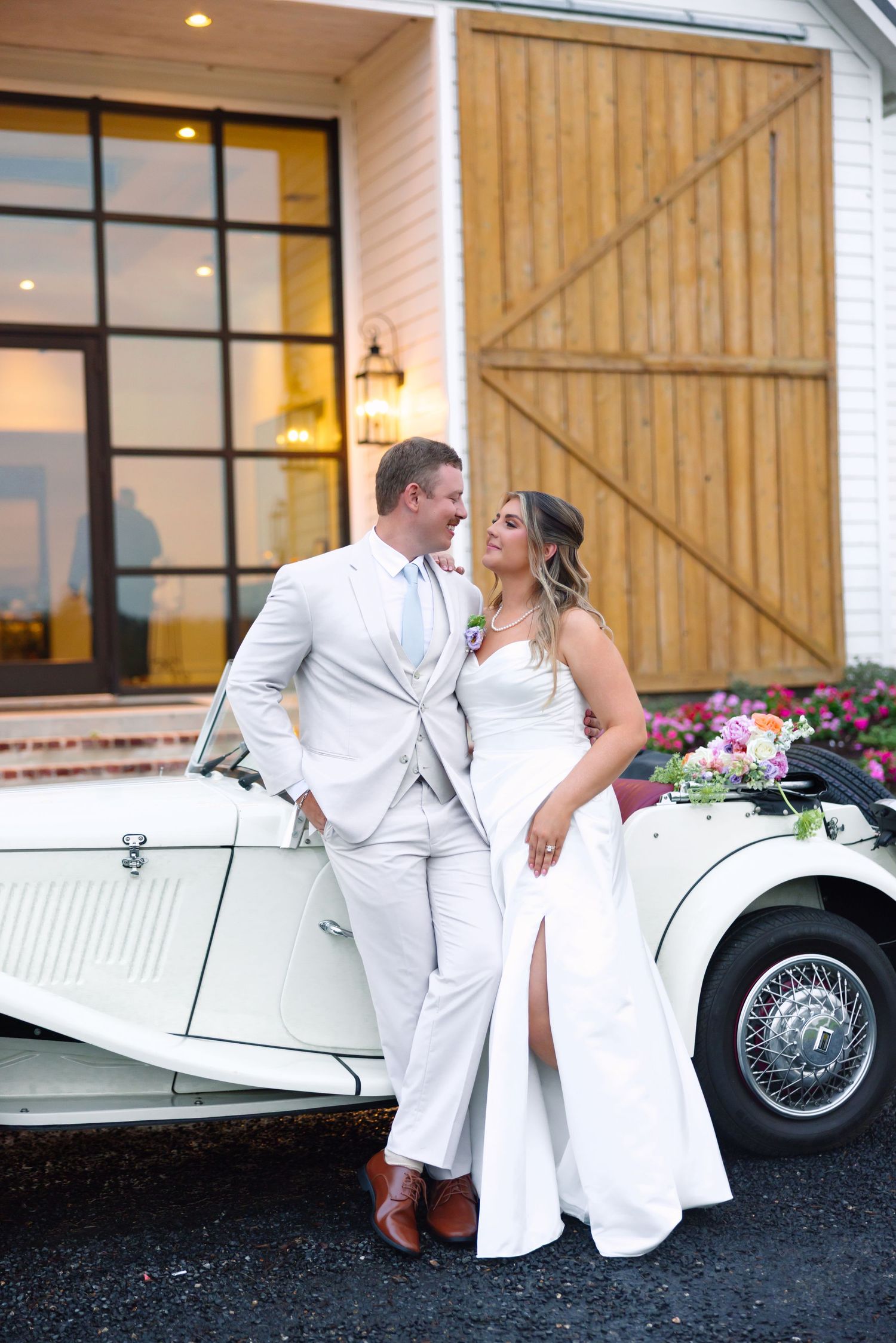 Bride and groom in white attire lean against a vintage white convertible car outside a barn venue.