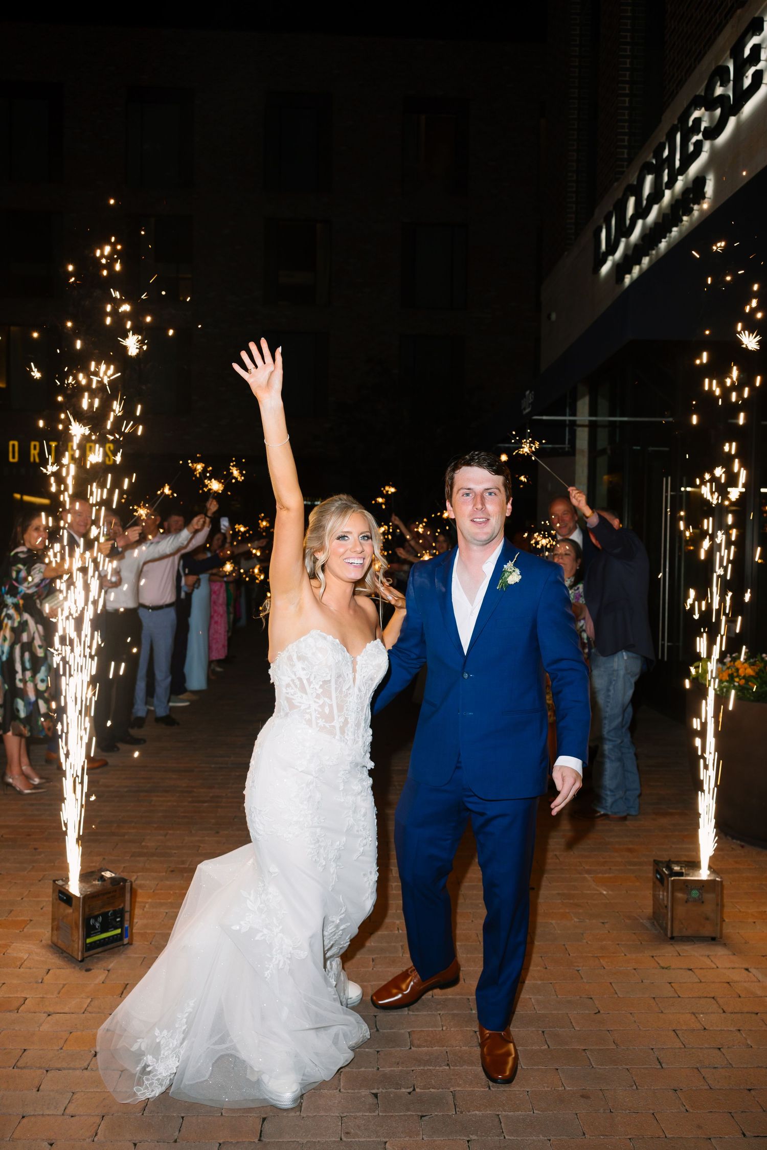 Bride and groom celebrate wedding exit with sparklers at night, bride raising arm joyfully in white gown and groom in blue suit.