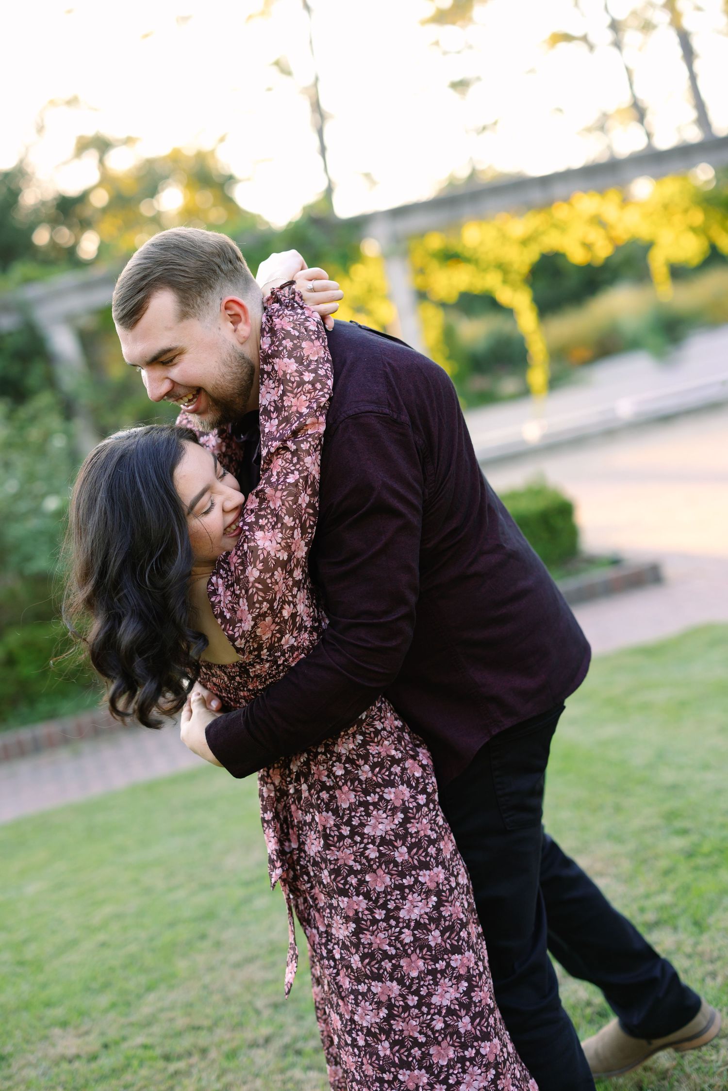 Couple embracing and laughing together in a lush garden at golden hour, woman in floral dress hugging man in burgundy shirt.
