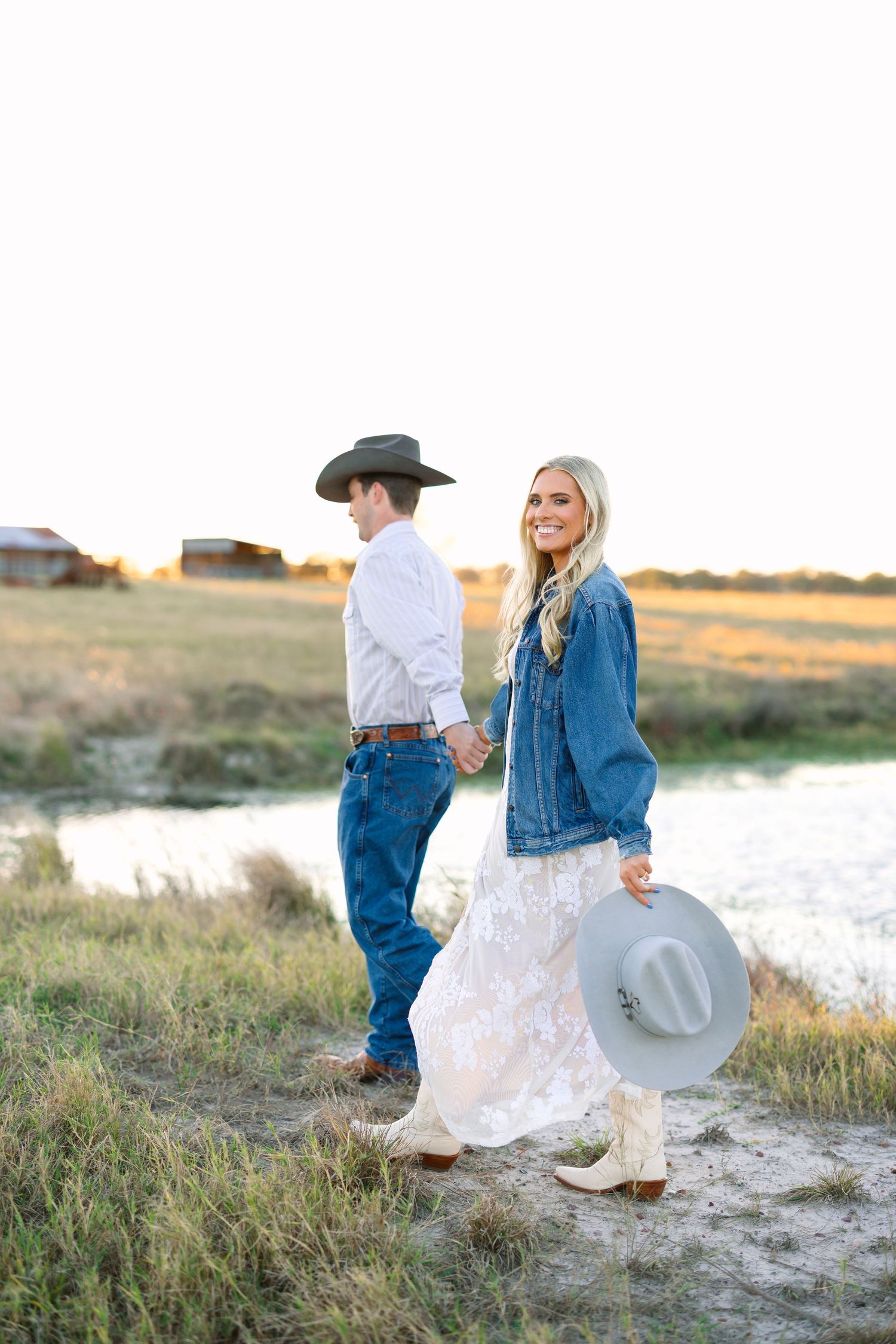 Blonde woman in denim jacket and lace dress holds a cowboy hat while walking hand-in-hand with her partner at sunset.