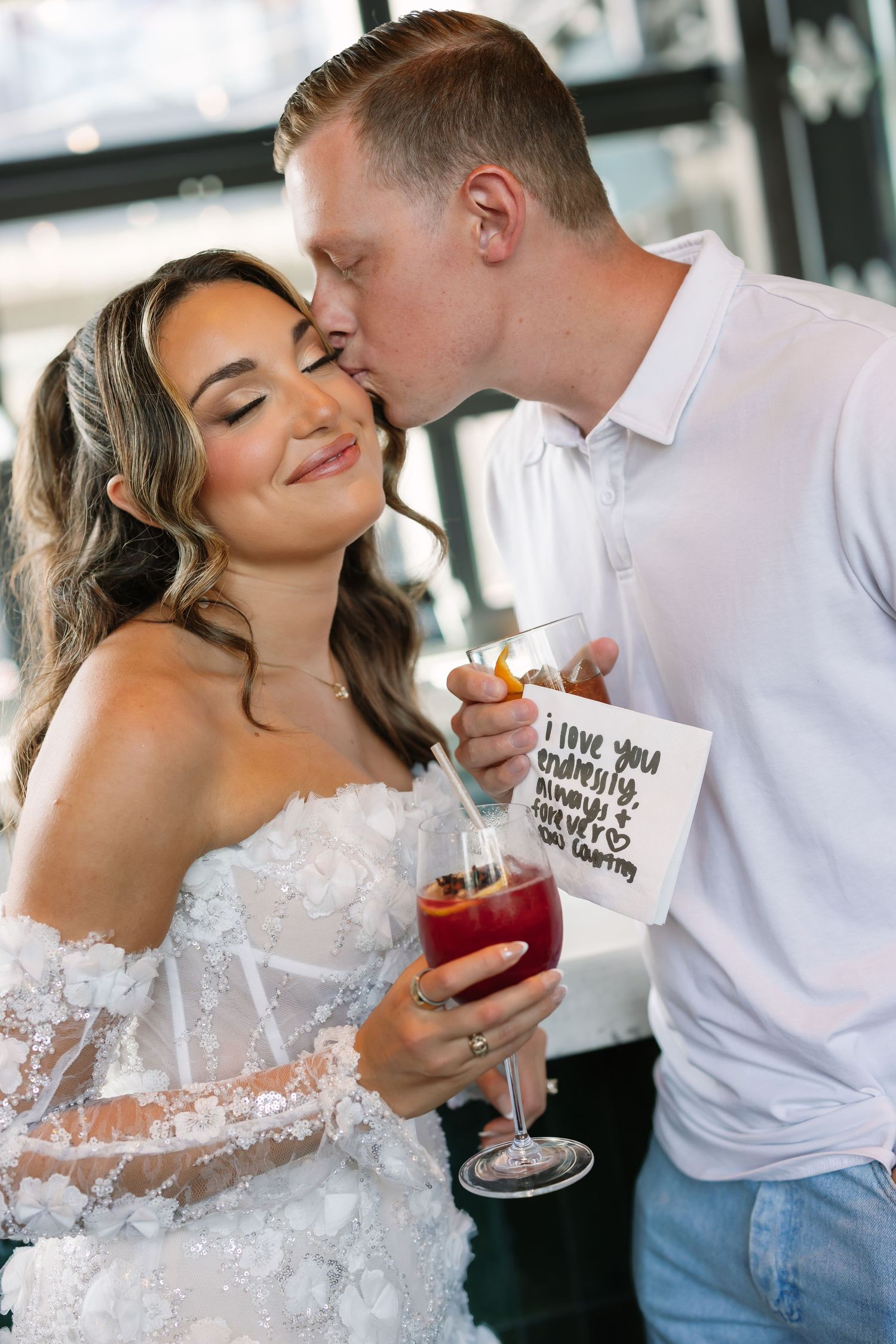 Groom kissing bride on cheek as she smiles holding a red cocktail and card at wedding reception.