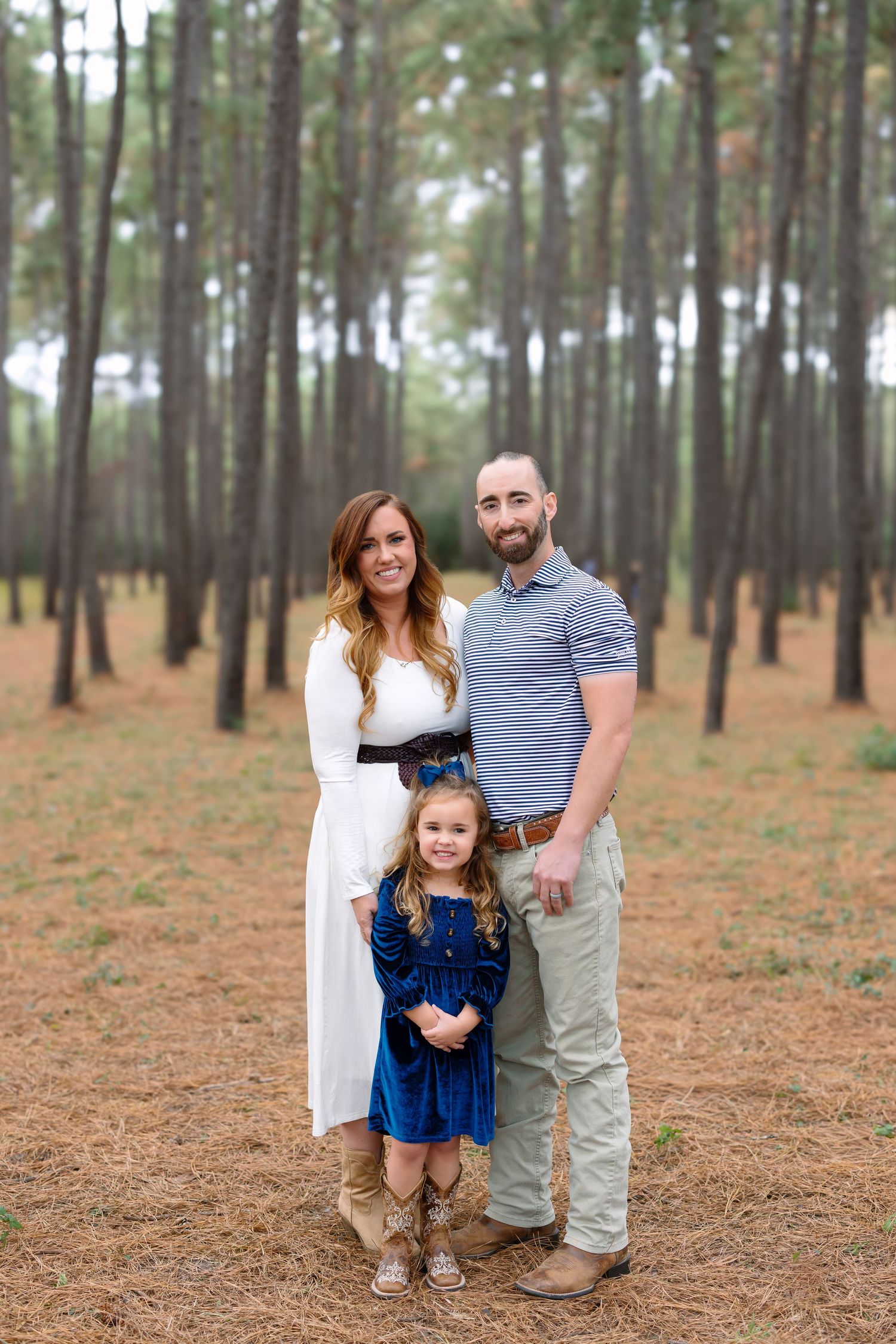 Family of three posing in a pine forest, woman in white dress, man in gray polo, young girl in blue velvet dress.