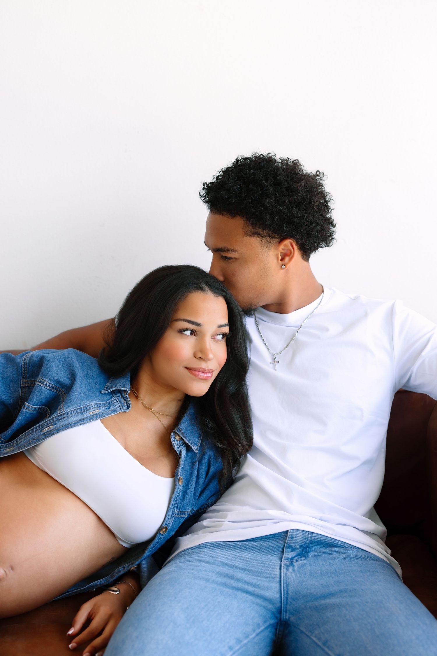A pregnant couple sitting together on a couch, woman leaning on man in white tee and jeans, both smiling softly.