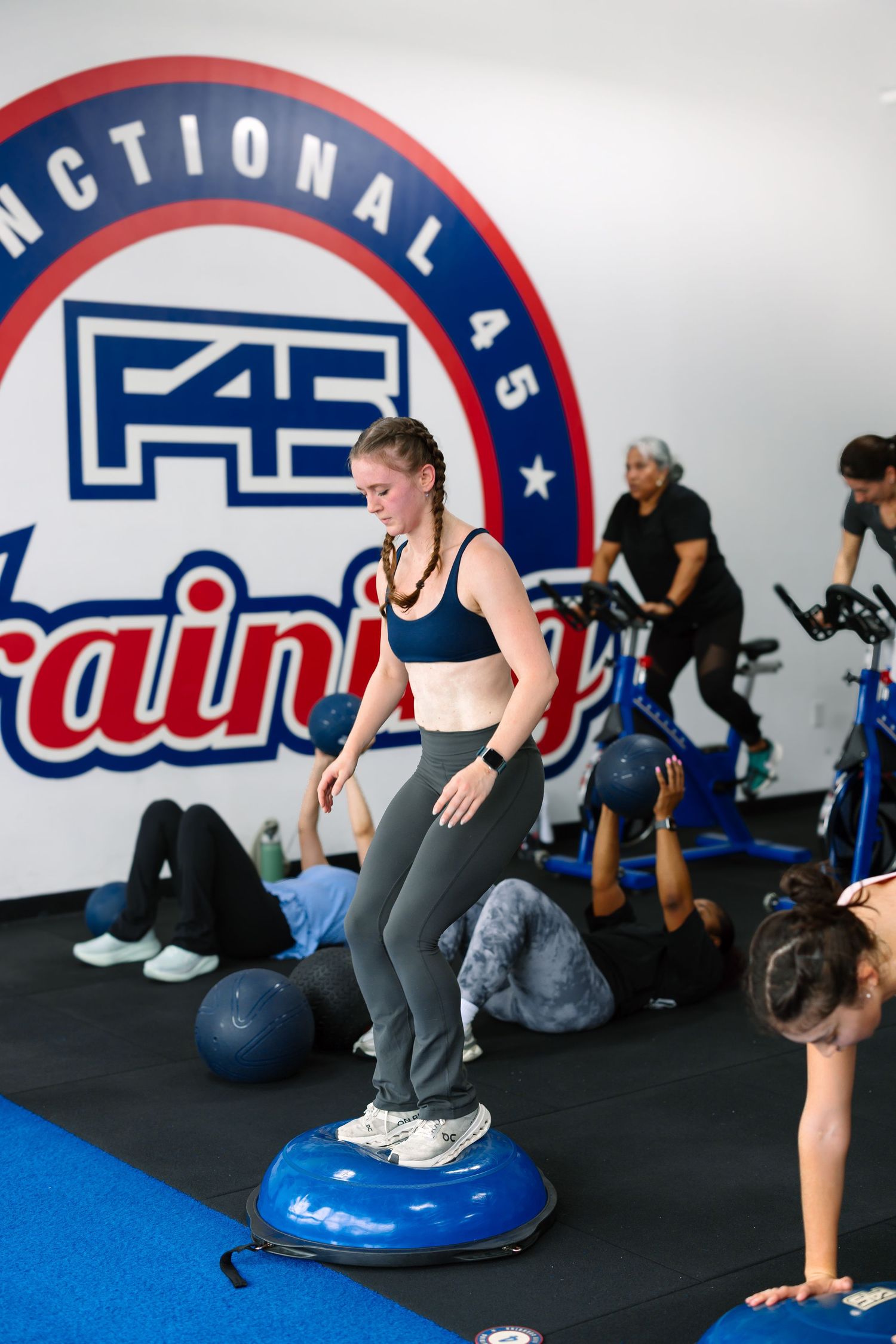 Woman balancing on a BOSU ball during an F45 functional training class with other members exercising in background.