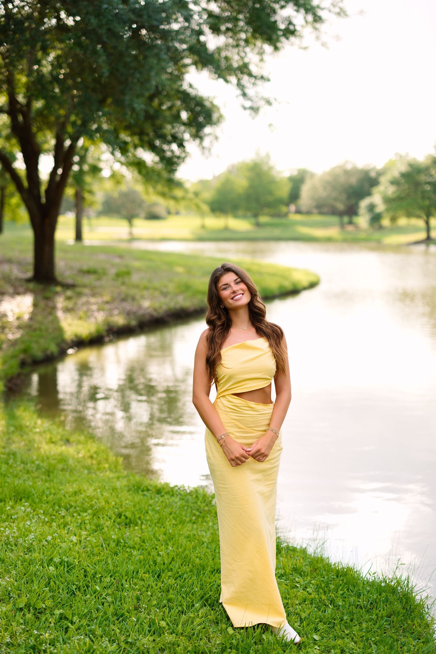 A person in a yellow formal dress poses by a serene lake during golden hour.