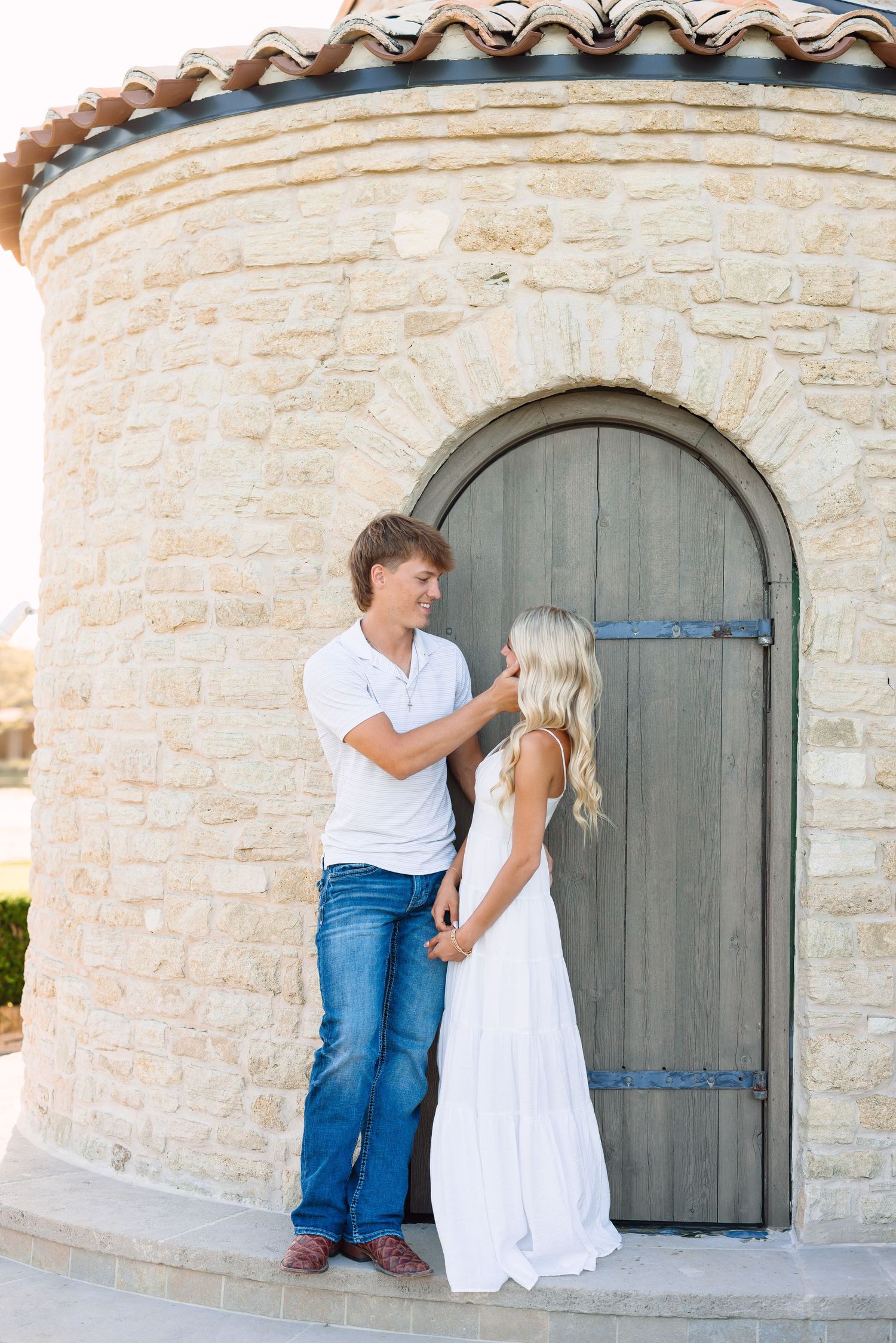 Couple poses playfully by stone building with arched wooden door.