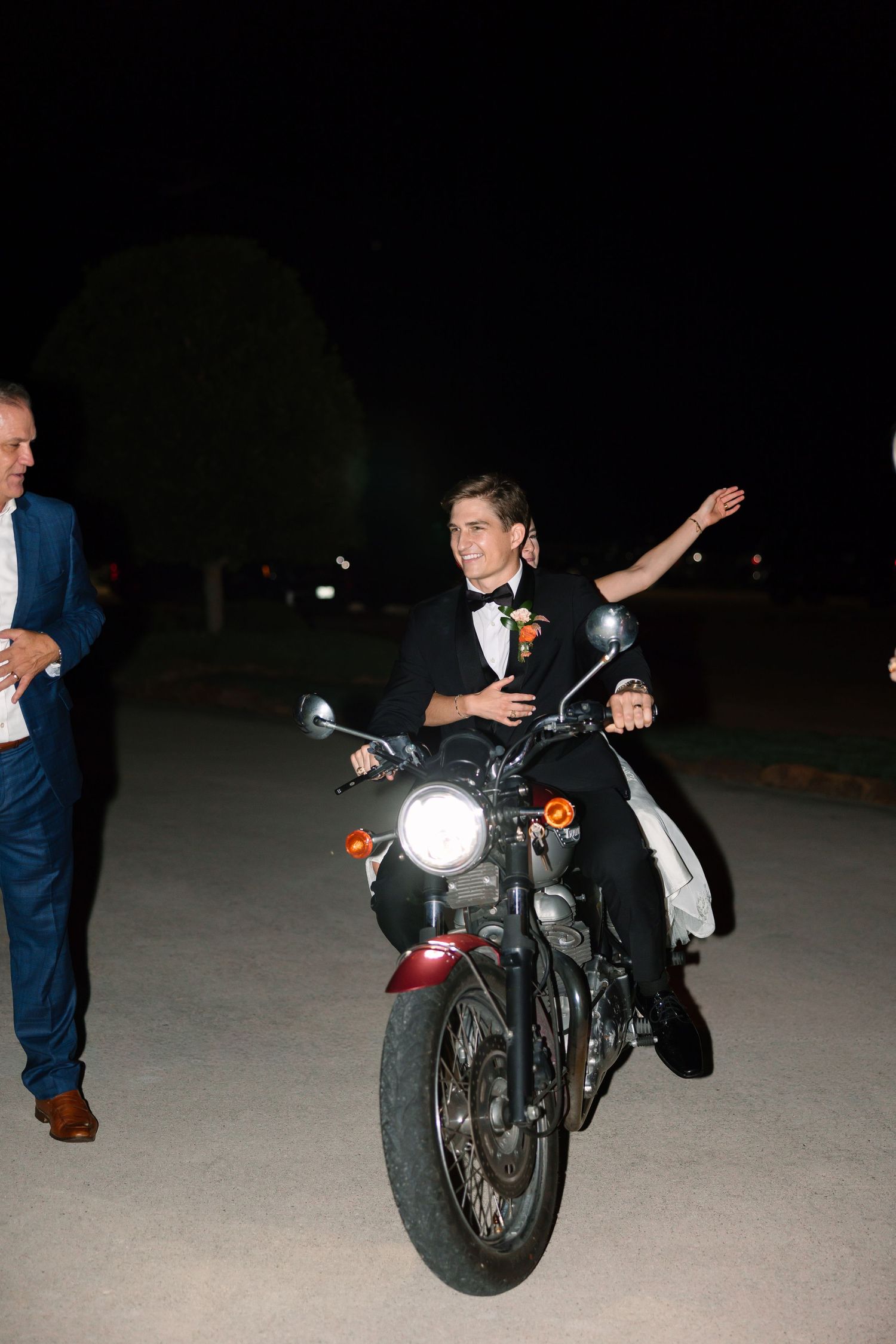 Groom in black tuxedo riding a motorcycle at night while waving, with guests standing nearby.