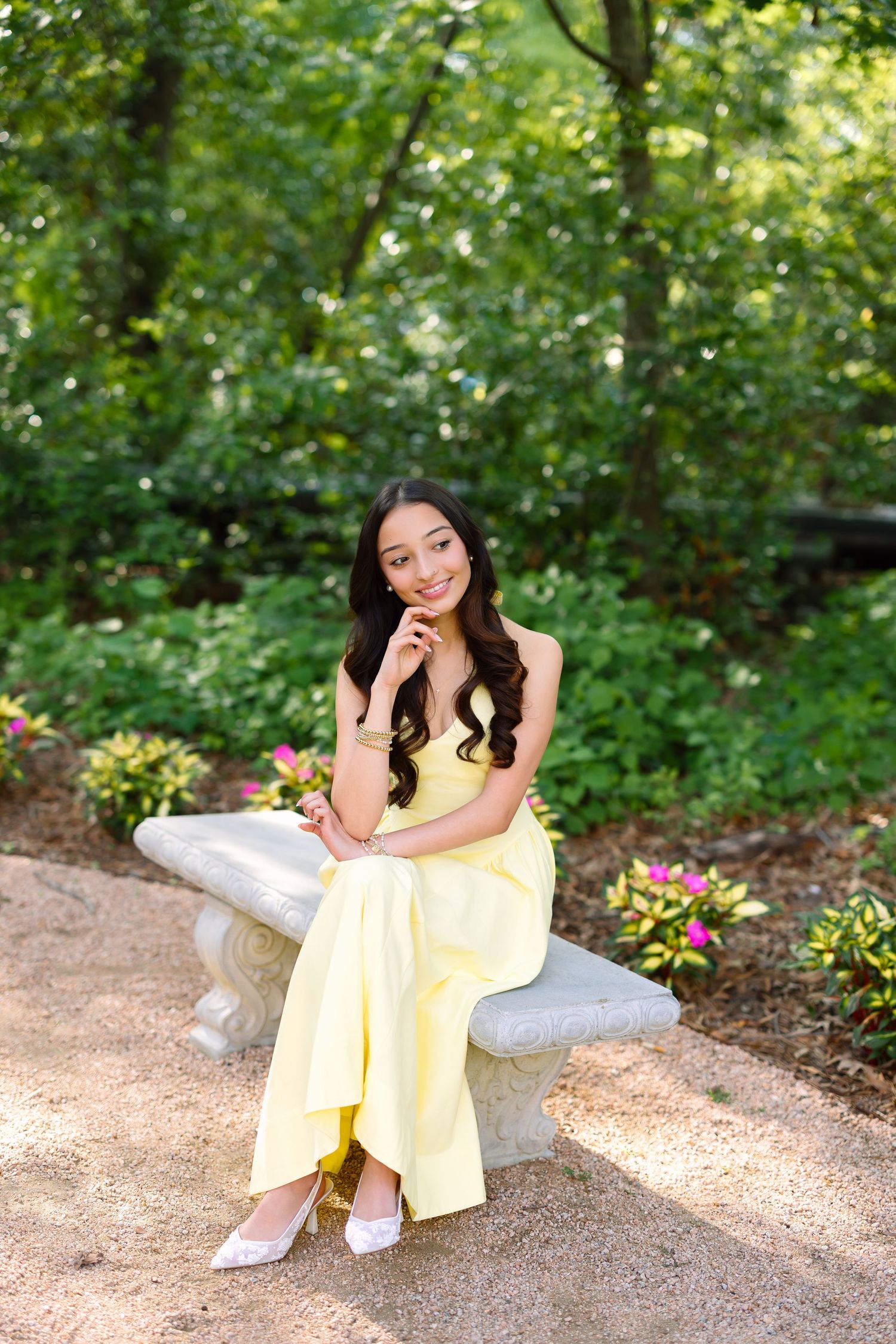 Young woman in yellow dress sitting on stone bench in lush garden, smiling gracefully for portrait photo.