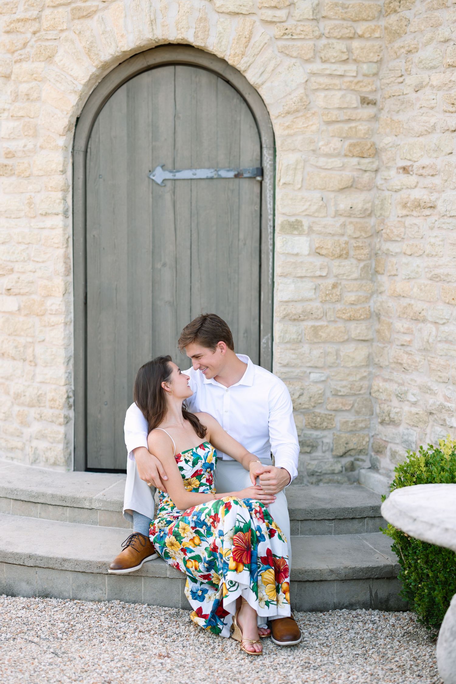 Couple sitting on stone steps in front of arched wooden door, woman in floral dress gazing at man in white shirt.