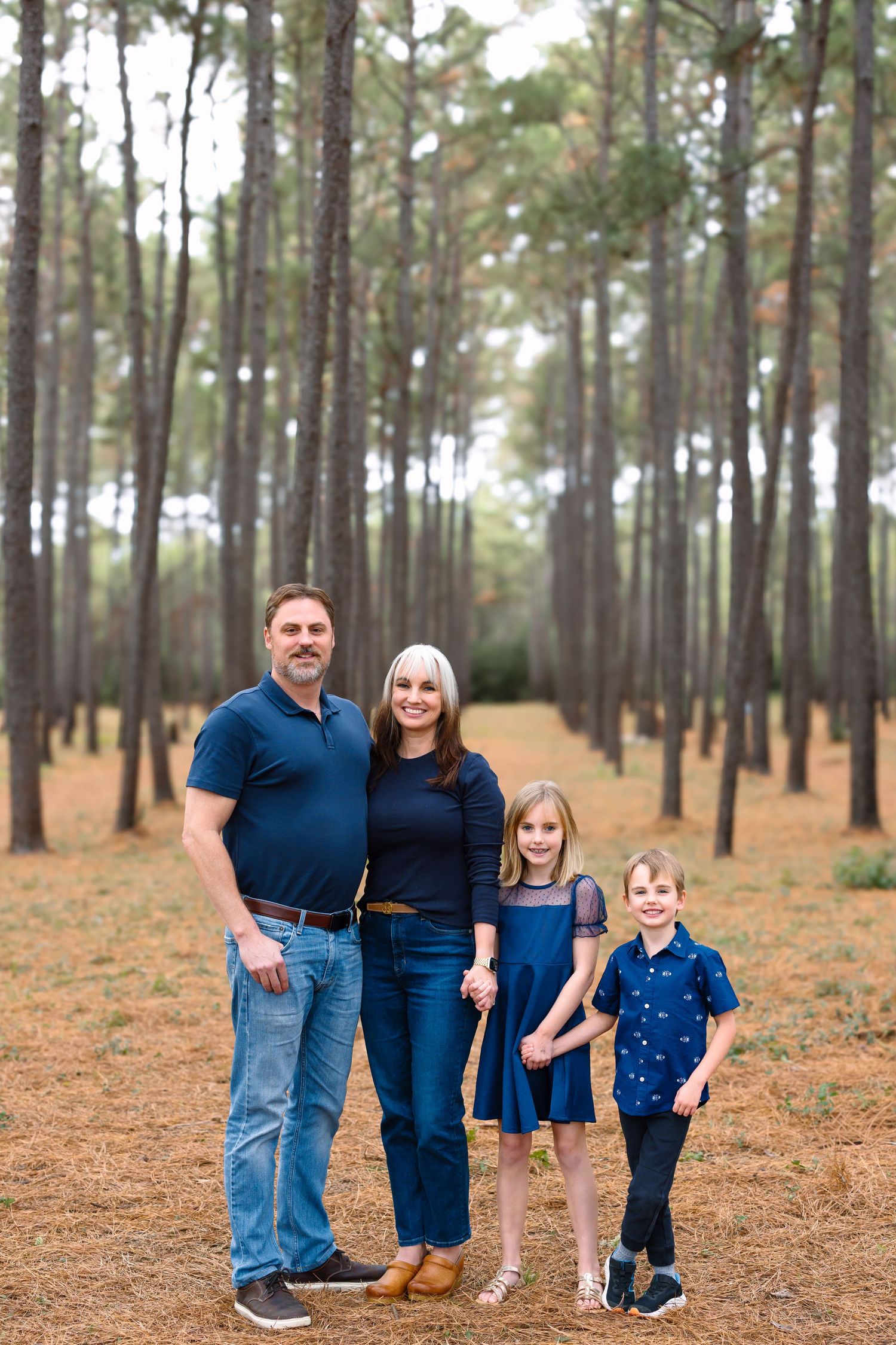 Family of four posing in a pine forest, wearing coordinating navy blue outfits for a professional outdoor portrait.