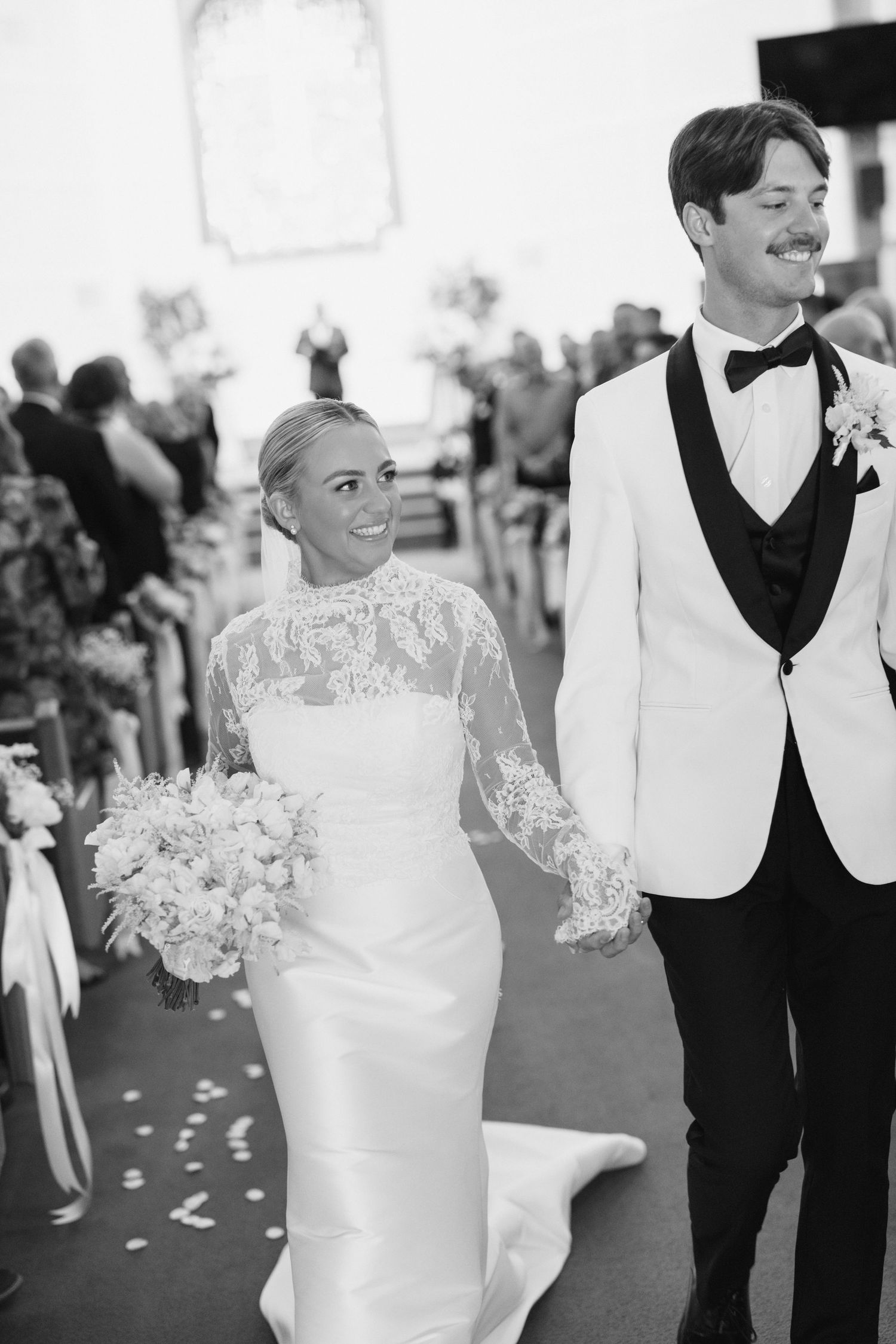 Black and white photo of a smiling bride holding bouquet and groom in white tuxedo walking down wedding aisle.