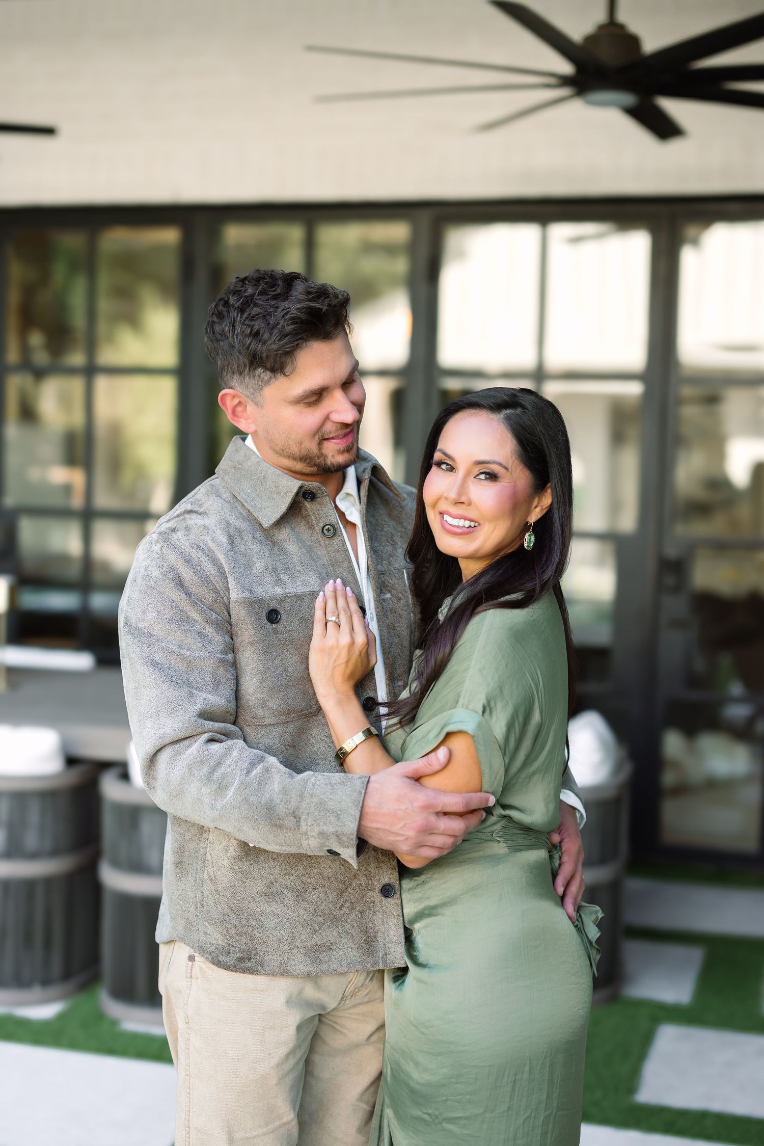A smiling couple embraces outdoors on a patio, woman in green dress and man in gray jacket, with a ceiling fan above.