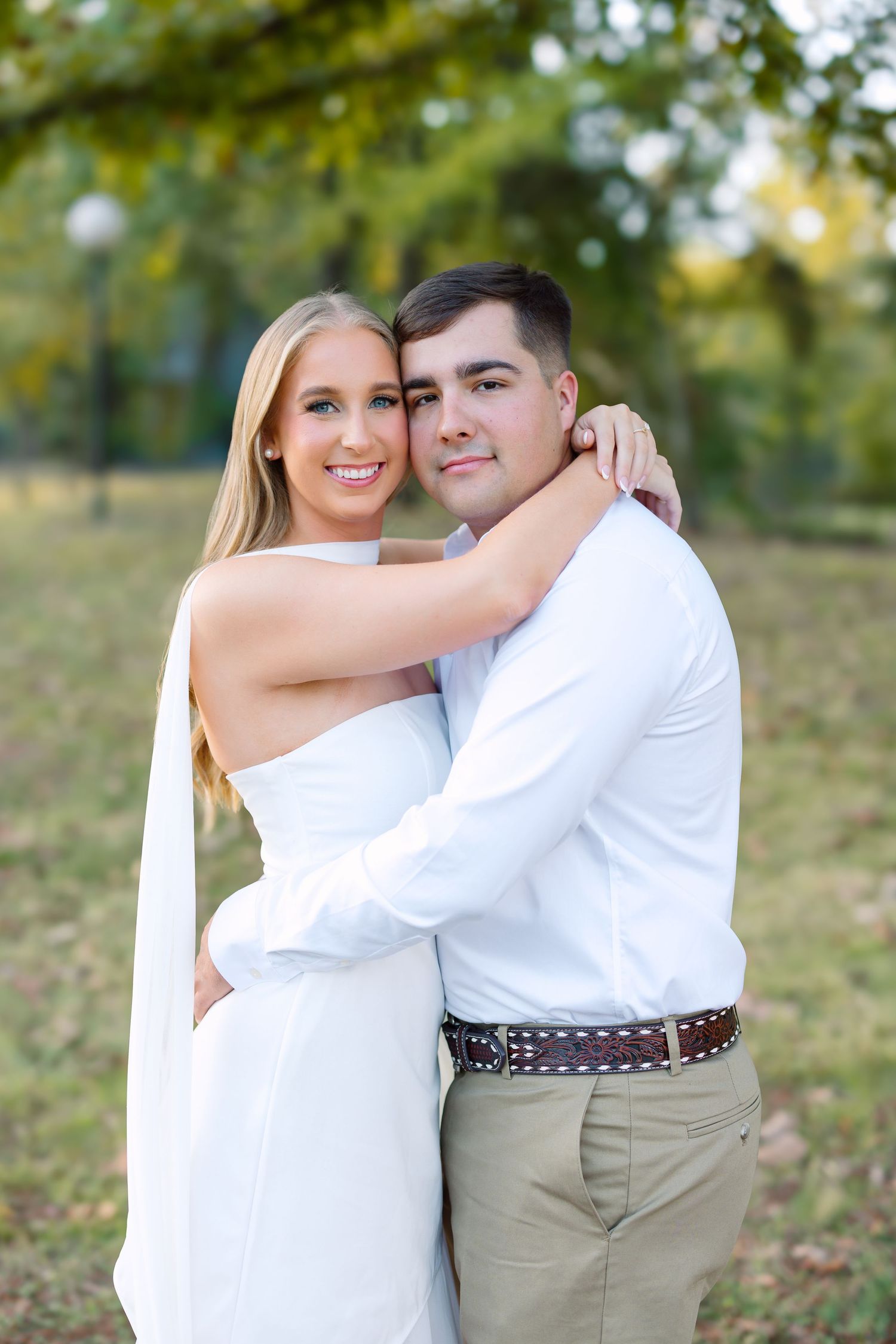 A smiling couple in white outfits embrace outdoors in a park, posing for an engagement or wedding photo.