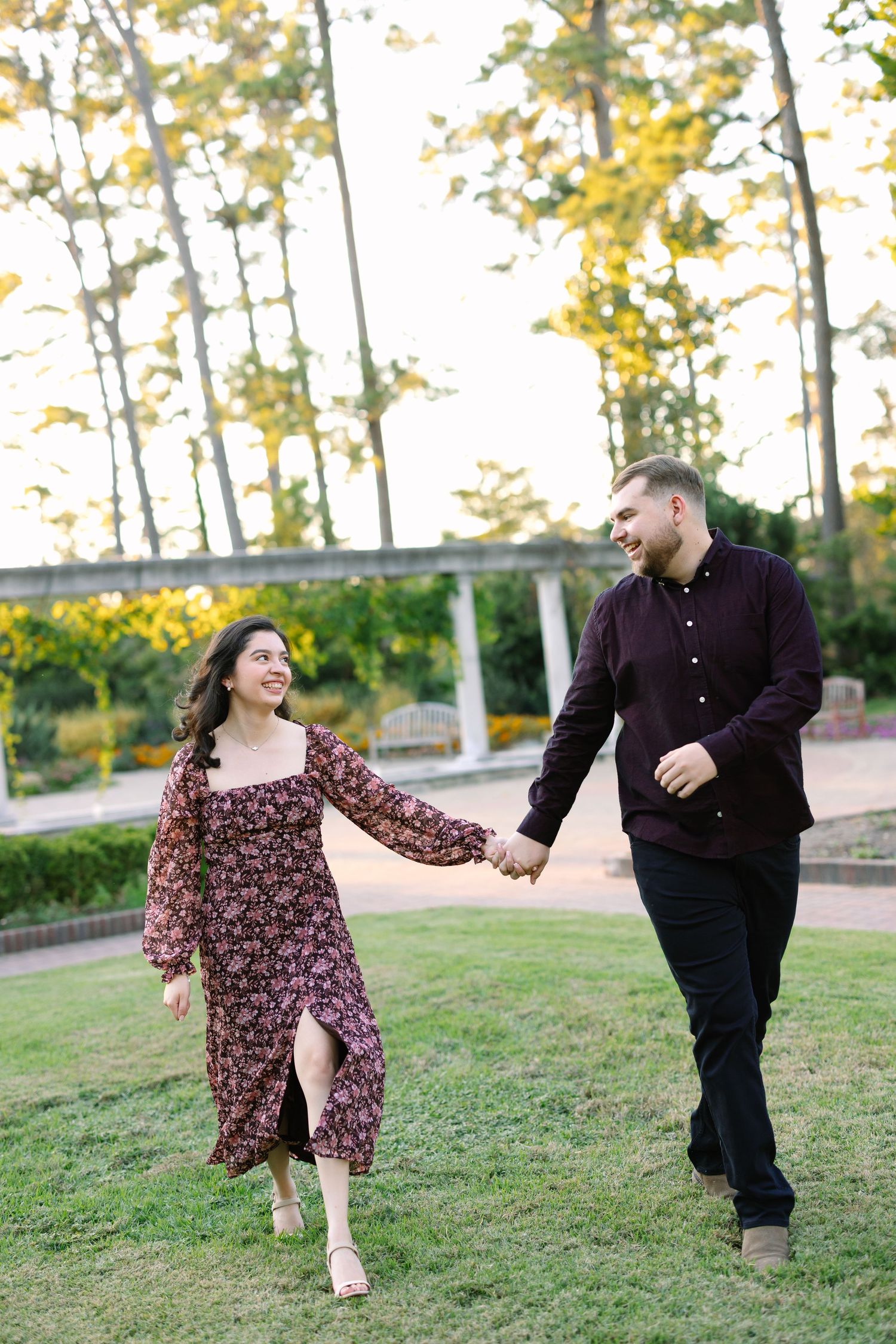 A couple holds hands and walks through a lush green park, smiling at each other, dressed in coordinating dark floral outfits.