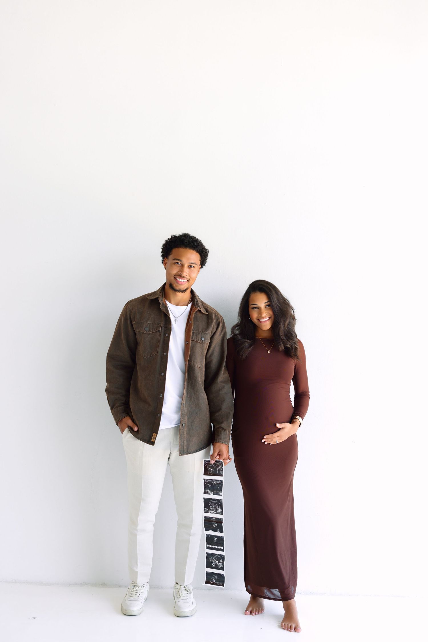 Couple posing against white wall, man in brown jacket, pregnant woman in burgundy dress holding ultrasound photos.