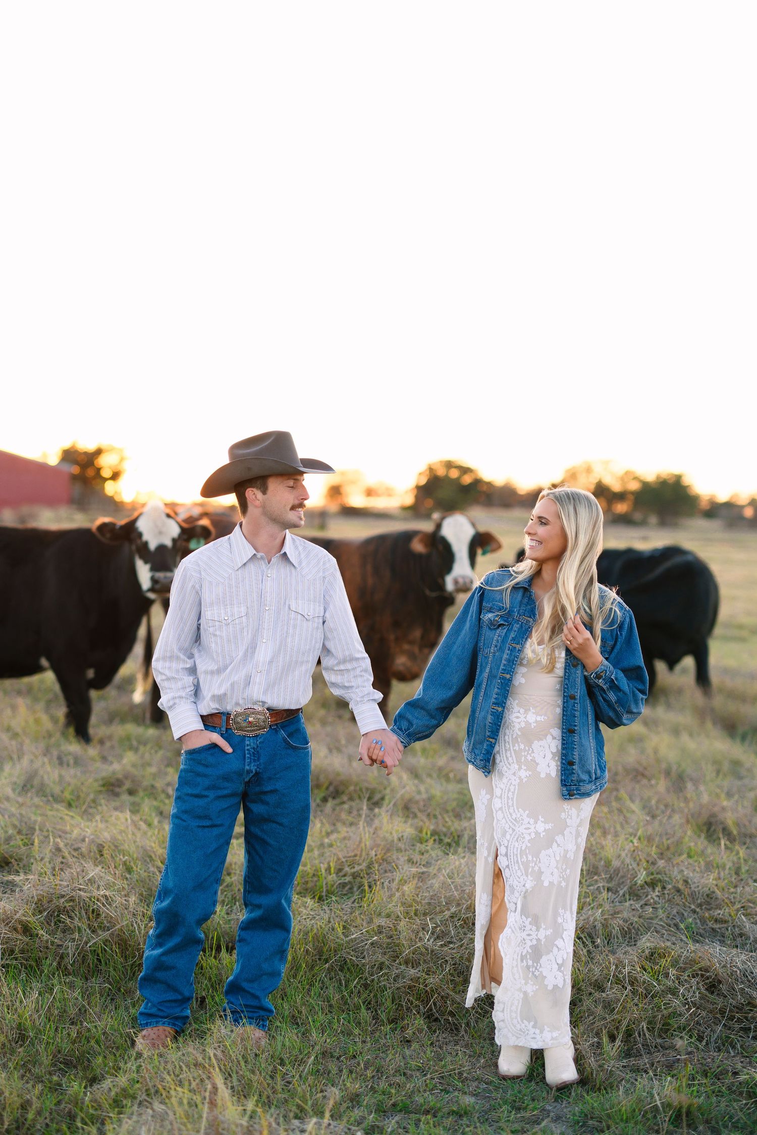 A couple holds hands in a sunlit pasture with black cattle, man in cowboy hat and jeans, woman in white dress and denim jacket.