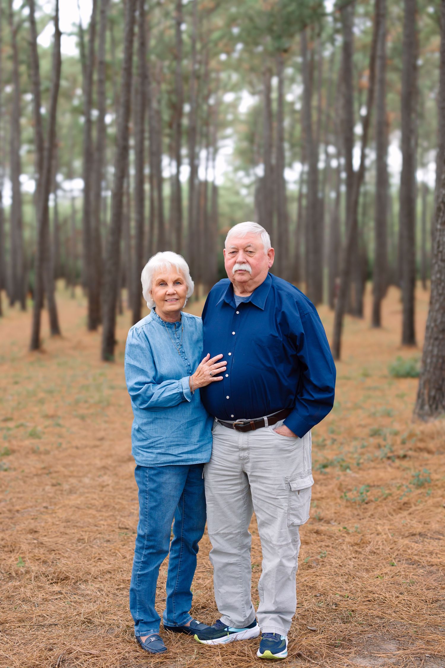 Elderly couple posing together in a pine forest, woman in denim jacket resting hand on mans chest.