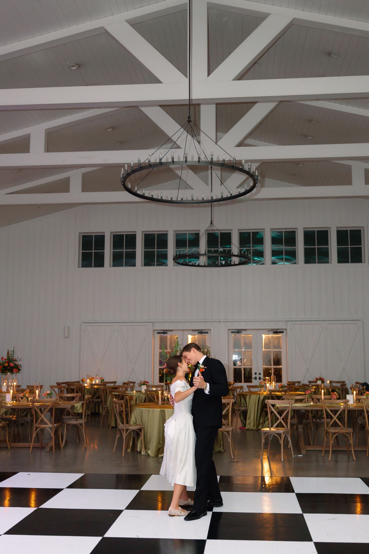 Bride and groom sharing first dance on black and white checkered floor in elegant white barn venue.