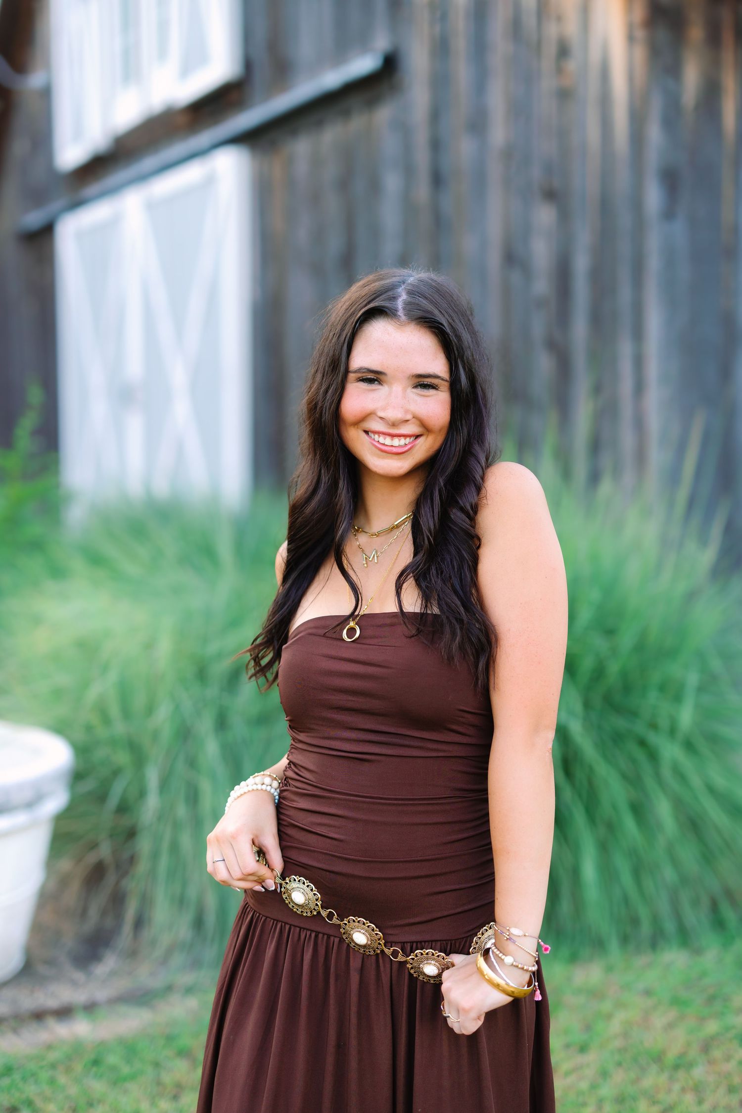 A brown summer dress with gold belt accessory is featured against weathered wooden walls.