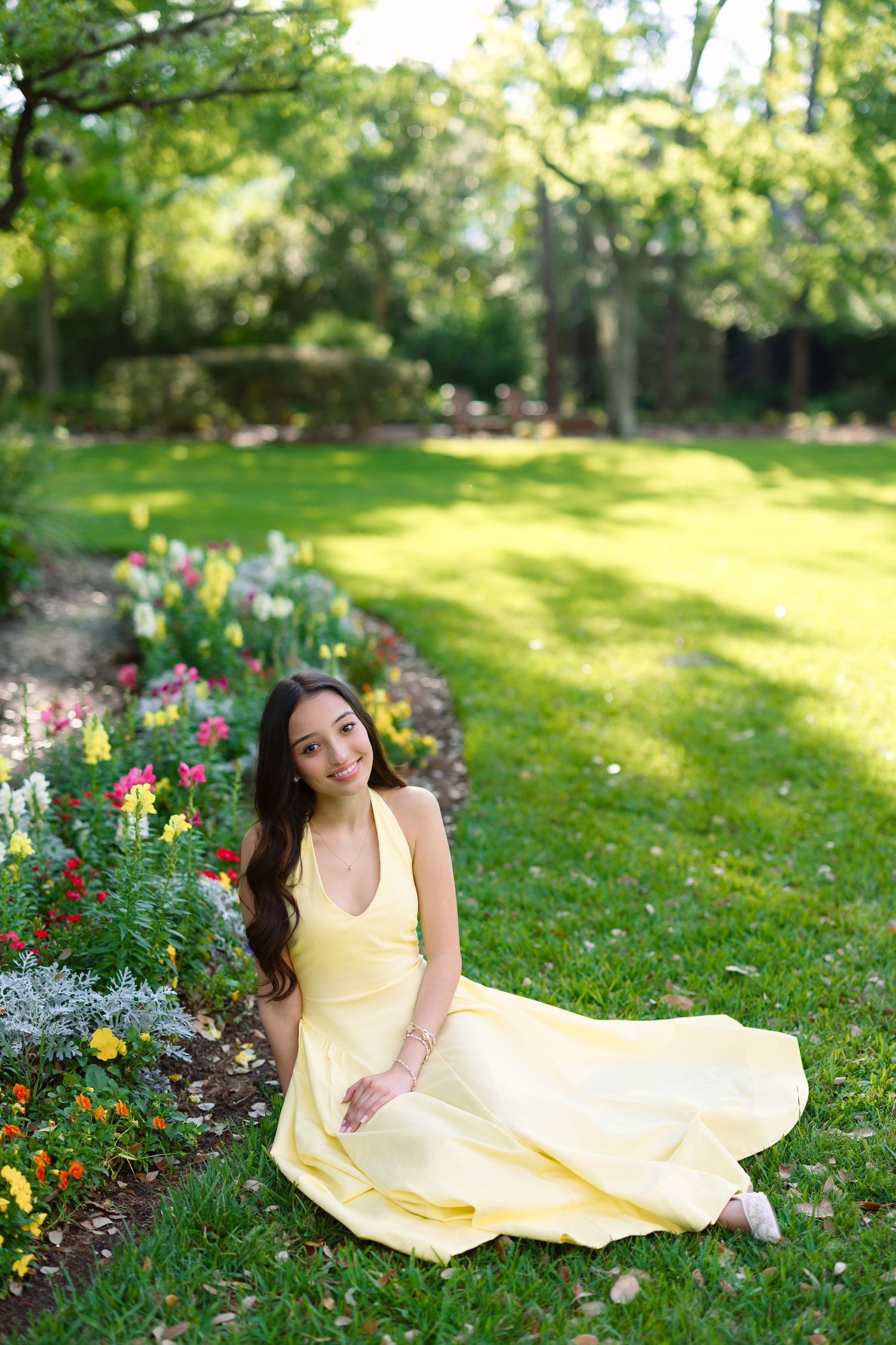 Young woman in yellow dress sitting gracefully on grass beside colorful garden flowers in a lush green park.