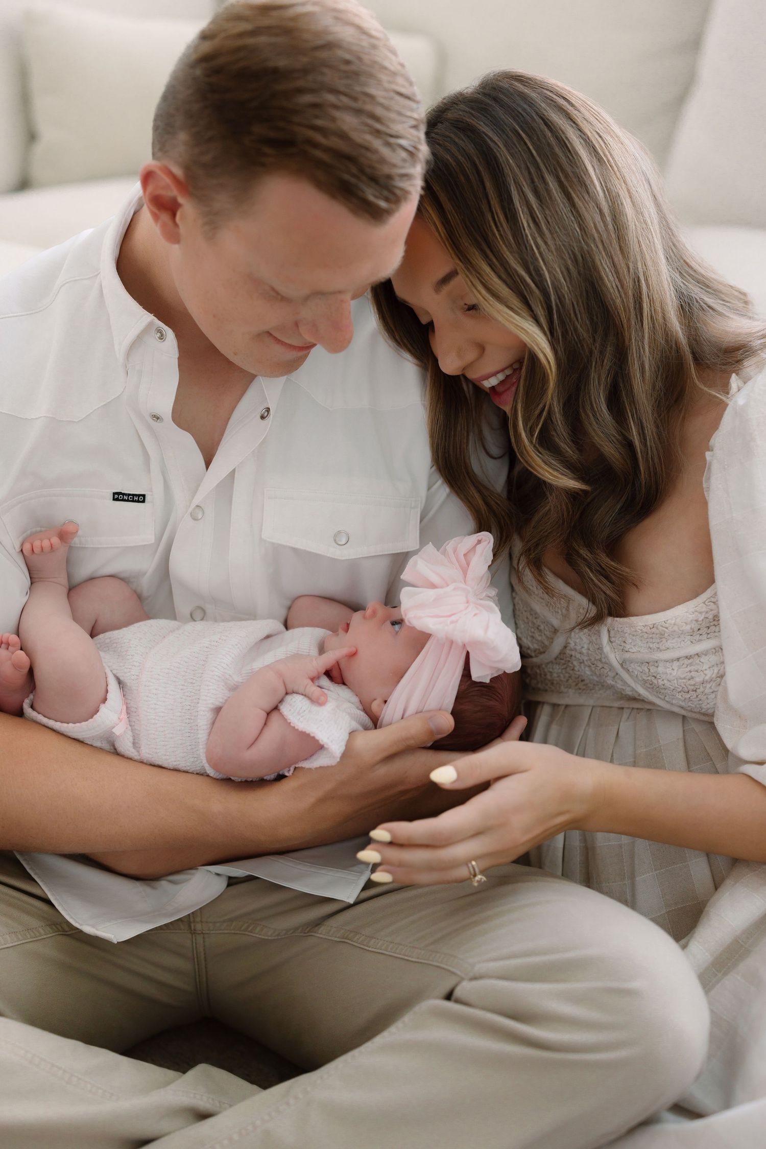 A couple smiles lovingly at their newborn baby dressed in pink, sitting together on a cream couch.