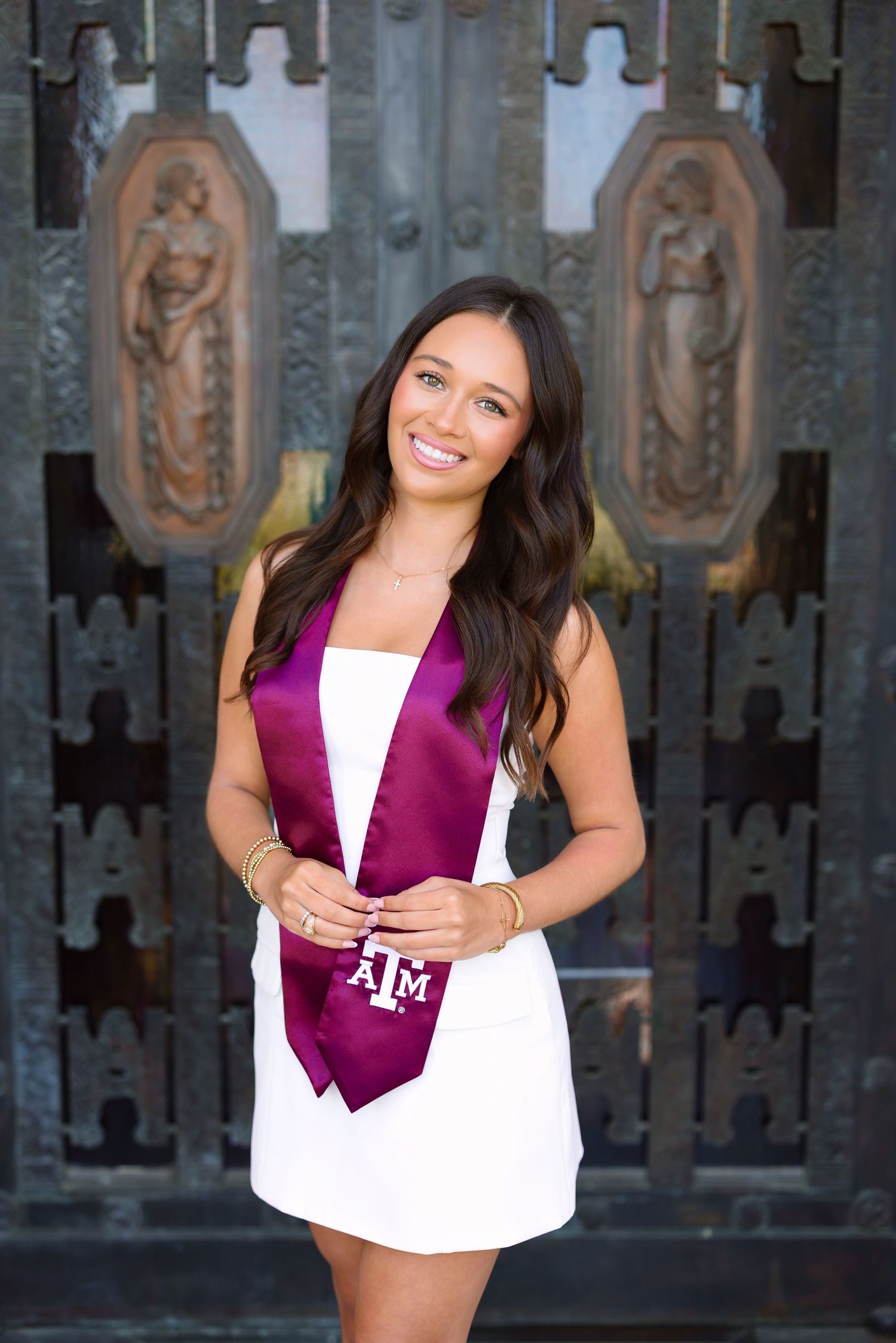 Smiling graduate in white dress wearing maroon Texas A&M sash poses before ornate bronze doors.