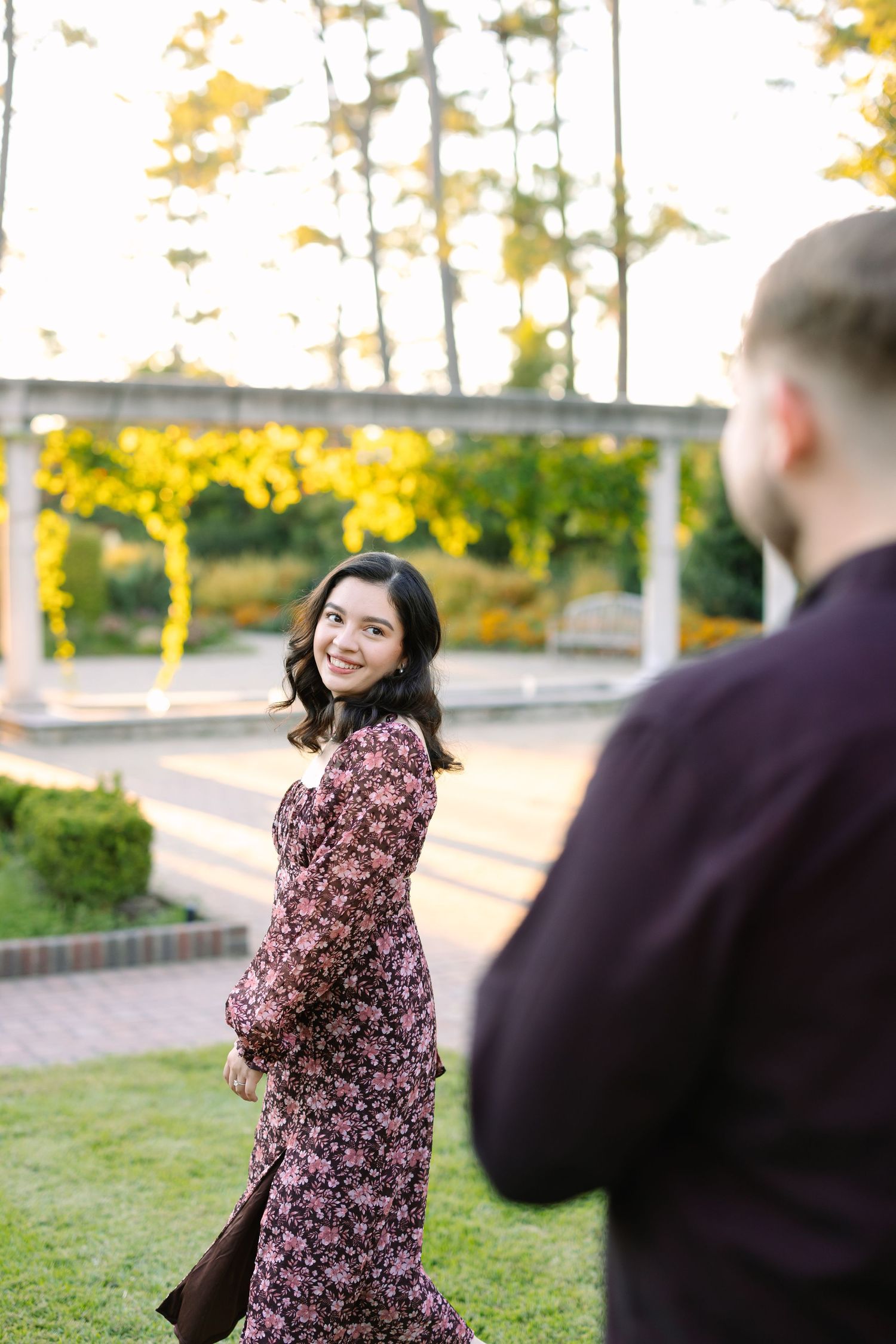 Woman in floral dress smiles over shoulder at man in garden setting with golden autumn trees.