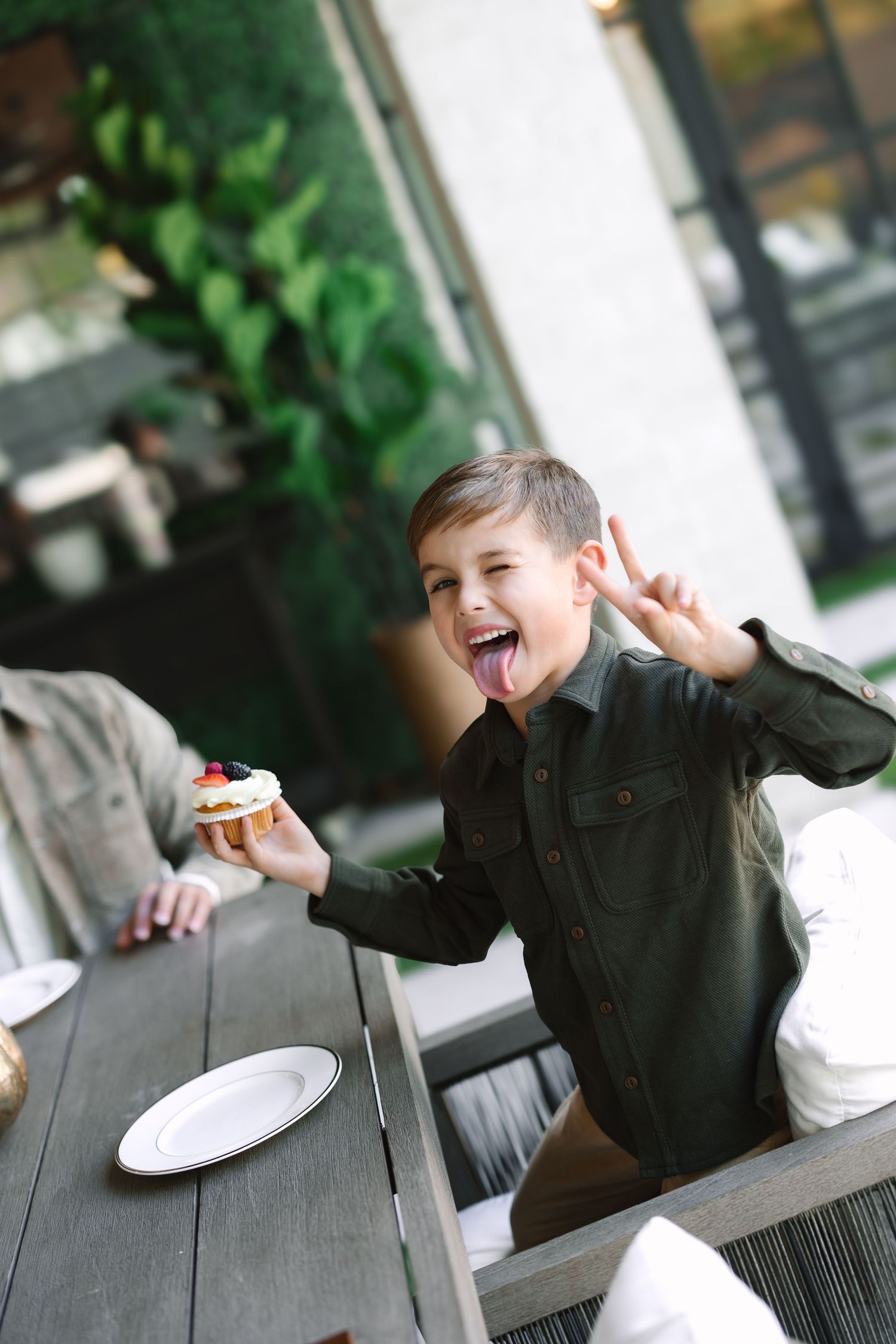Excited boy sticking tongue out, making peace sign, holding a cupcake at an outdoor restaurant table.