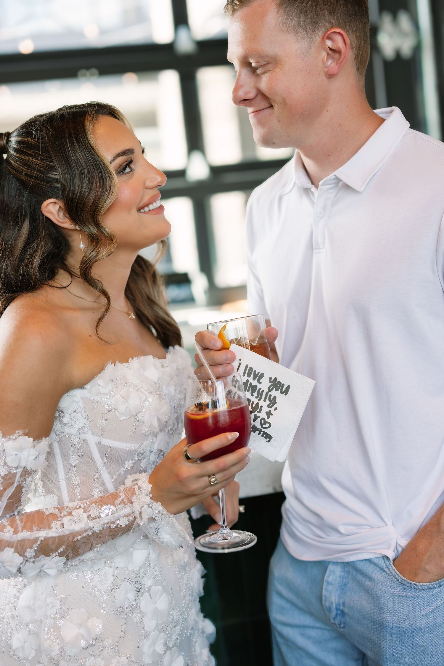 Bride in floral white gown smiling at groom in white polo, both holding red cocktails at wedding reception.