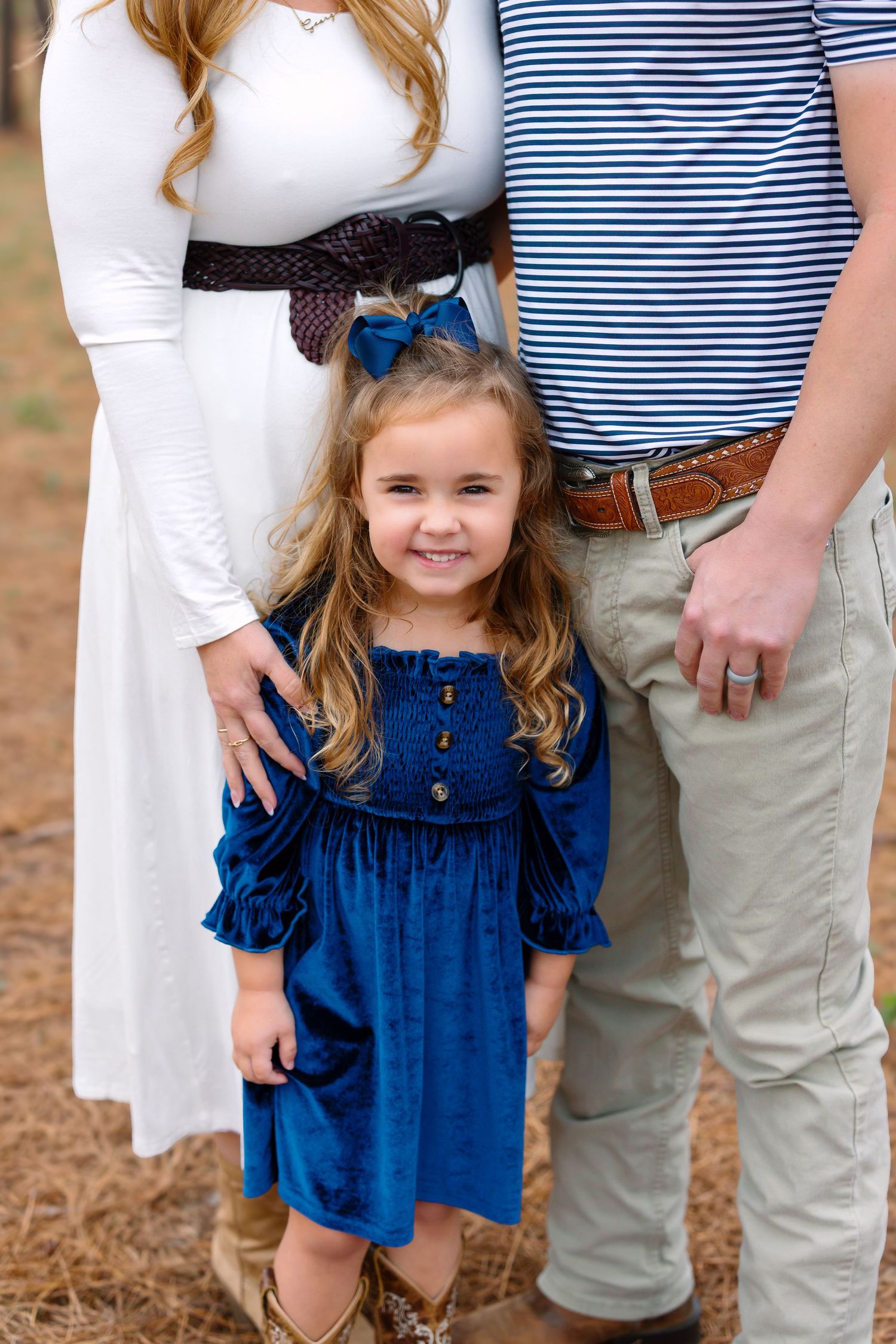 A smiling little girl in a blue dress stands between her parents during a family portrait session outdoors.