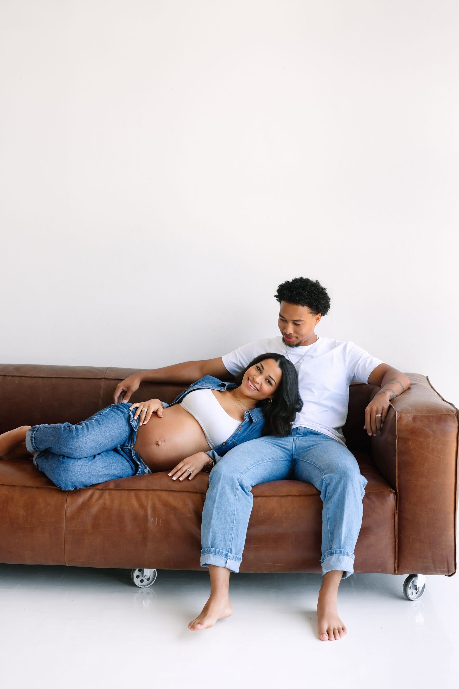 A pregnant couple relaxing on a brown leather sofa, woman lying with head on partner's lap, both smiling warmly.