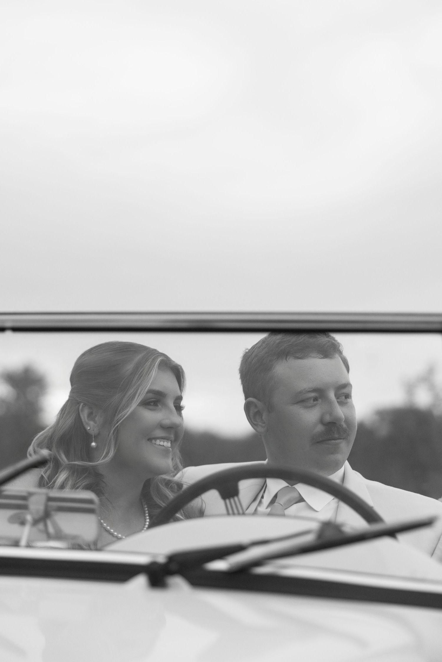 Black and white photo of a smiling bride and groom sitting in a vintage convertible car on their wedding day.