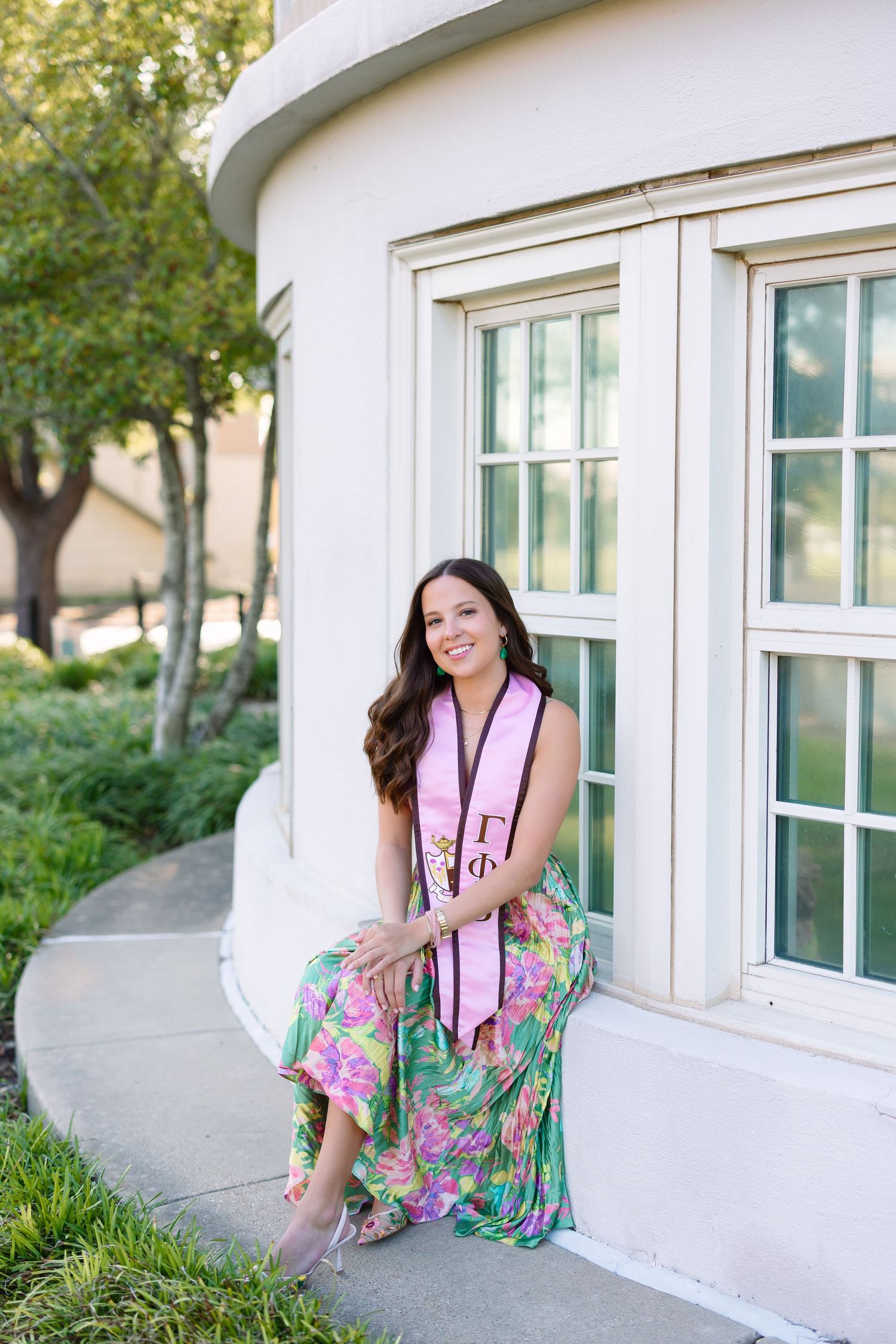 A floral dress and pink graduation stole create a cheerful outdoor portrait against a white curved building wall.