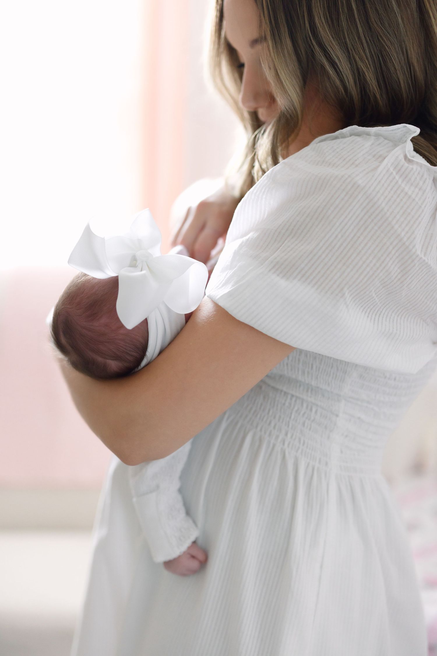 Mother in white dress cradling newborn baby with white bow in softly lit nursery room.