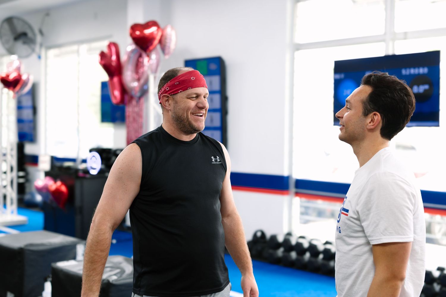 Two men smiling and talking in a gym with red heart balloons, weights, and blue flooring in the background.