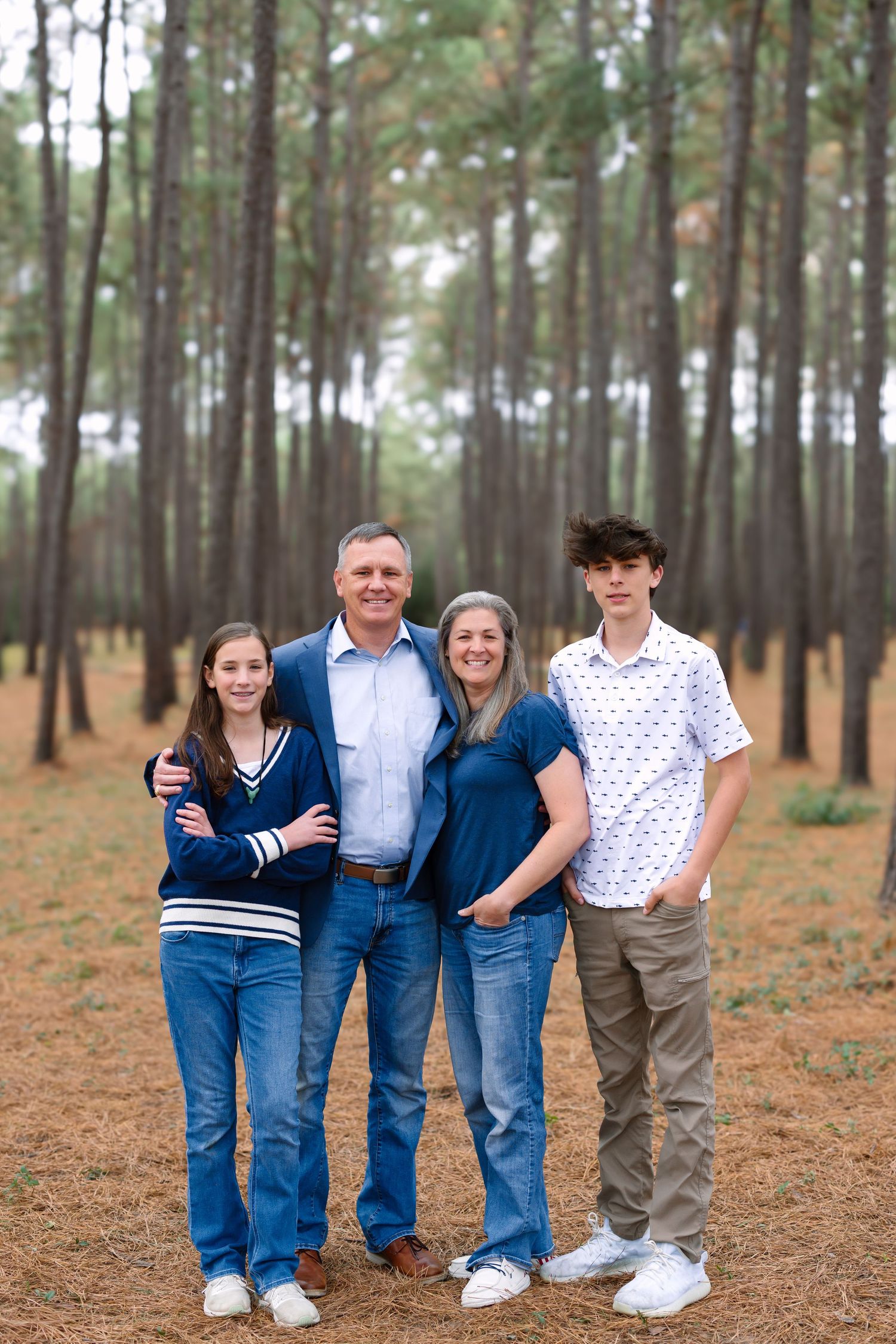 Family of four posing in a pine forest, wearing blue and casual attire, smiling together outdoors.