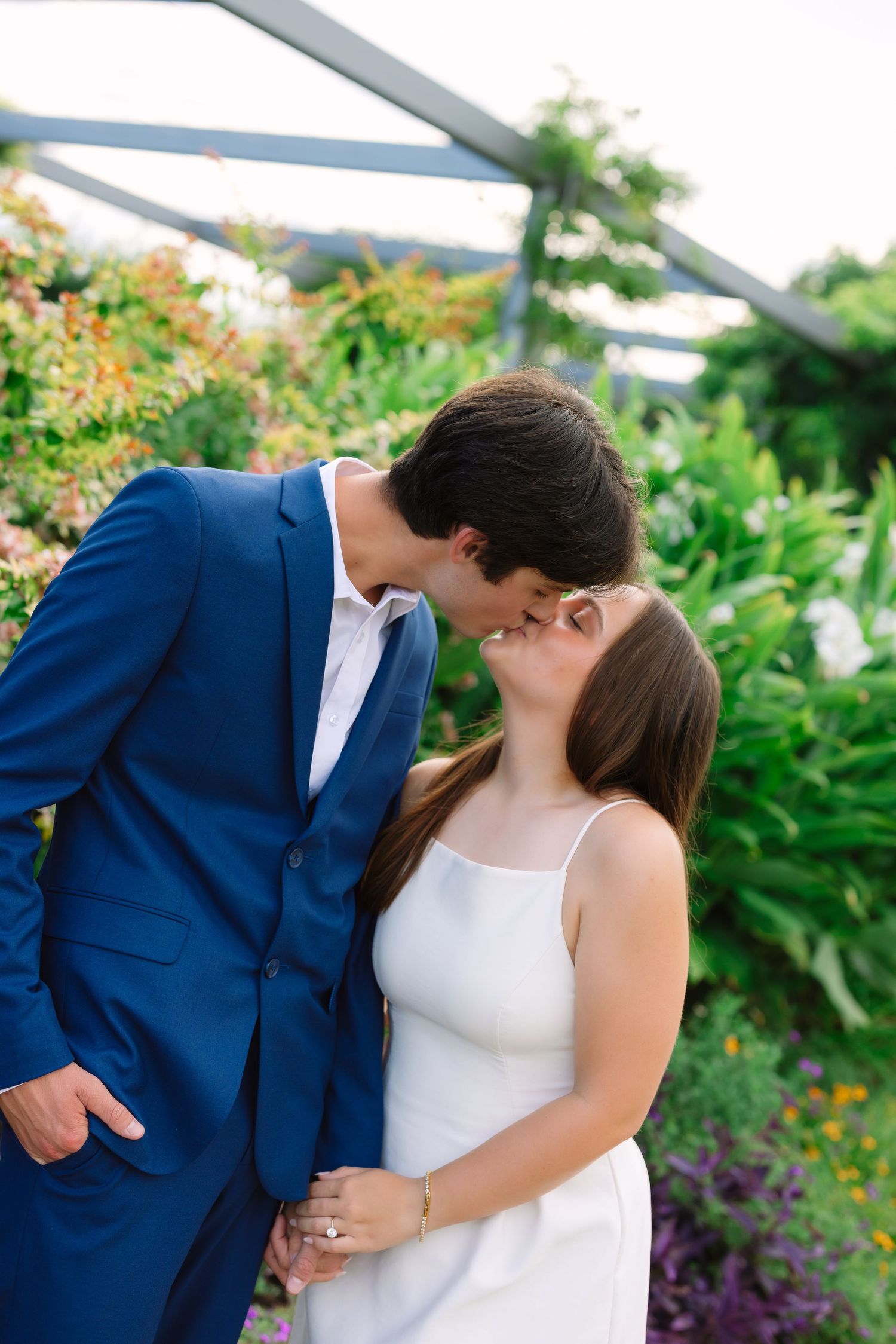 A couple shares romantic kisses in a garden setting with colorful flowers in the background.