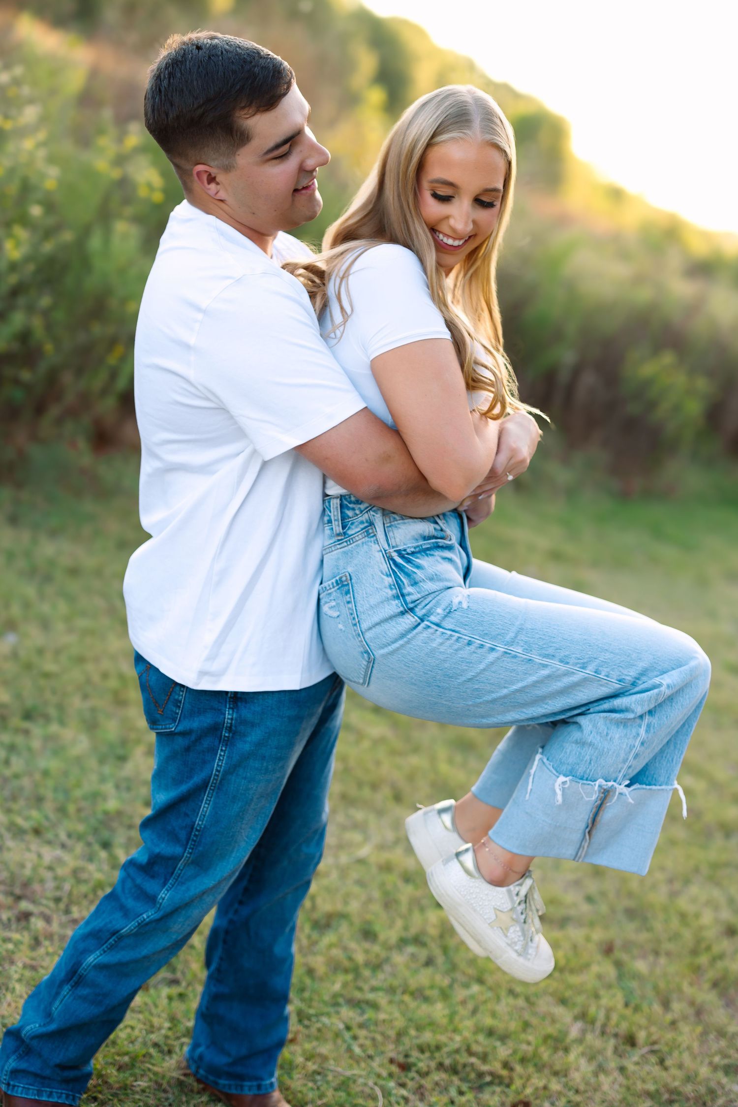 Man in white t-shirt and jeans playfully lifting smiling blonde woman in casual outfit in a sunny green field.