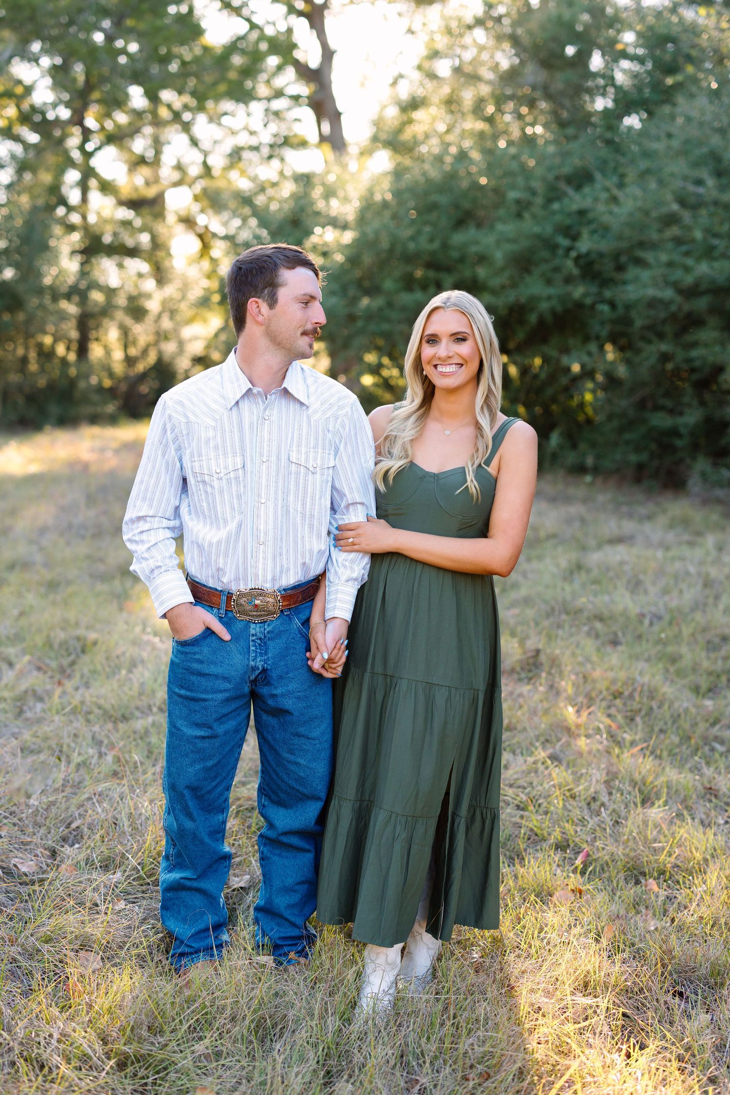 Couple holding hands in a sunlit field, man in jeans and striped shirt, woman in green maxi dress.