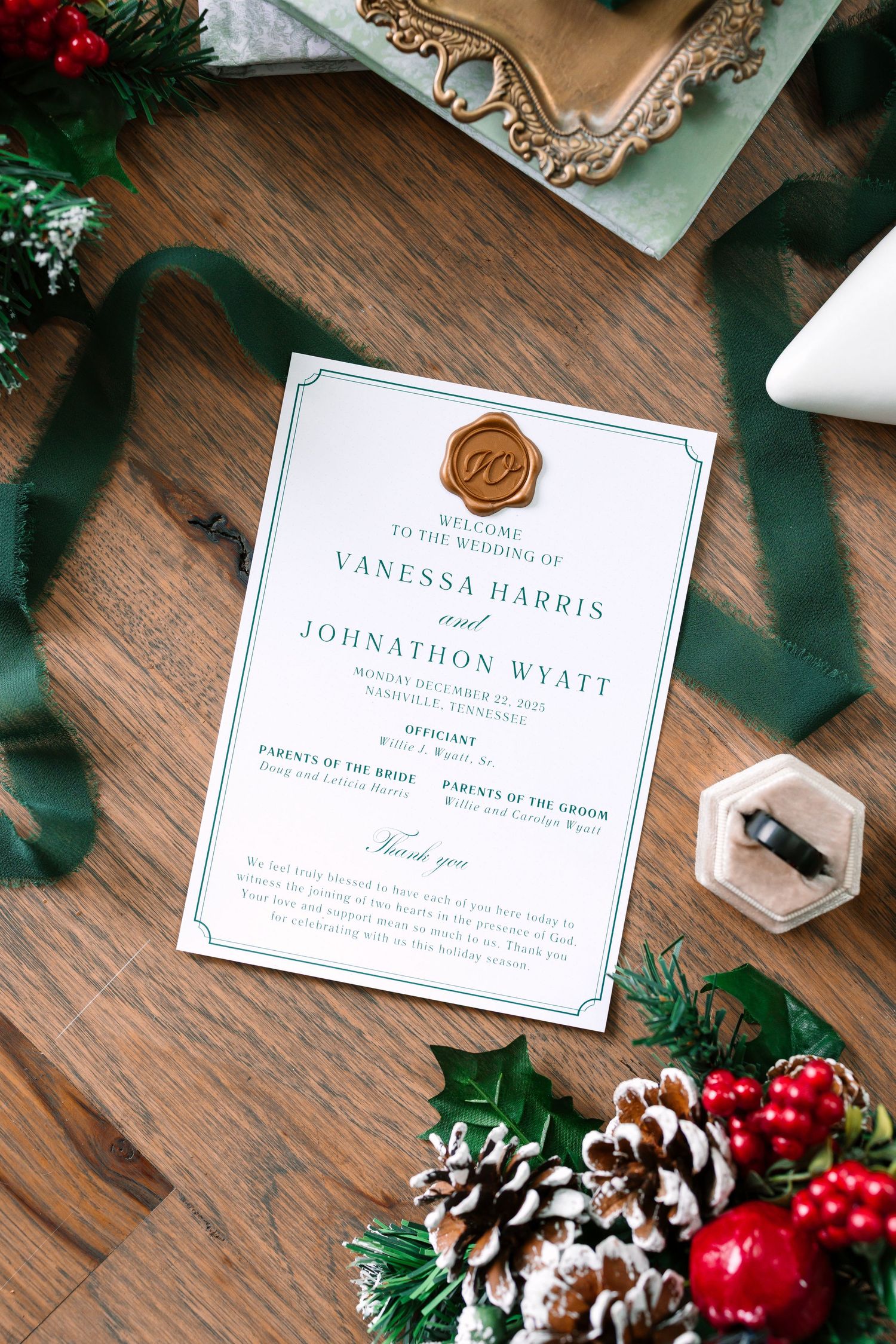Christmas wedding invitation for Vanessa Harris and Johnathon Wyatt with gold wax seal, green ribbon, holly, and pinecones on wood.