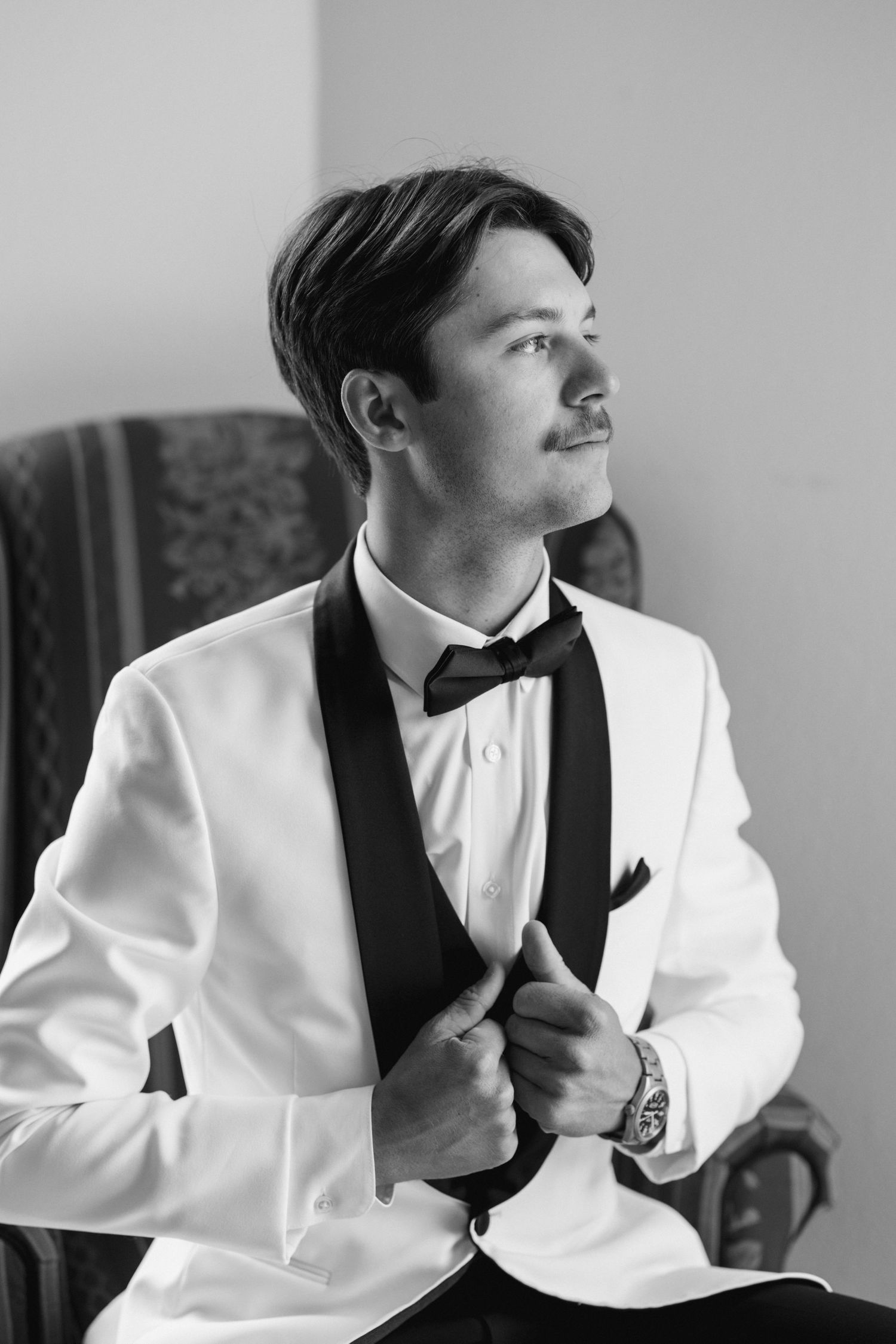 Black and white portrait of a dapper groom in white tuxedo and bow tie adjusting his jacket while seated.