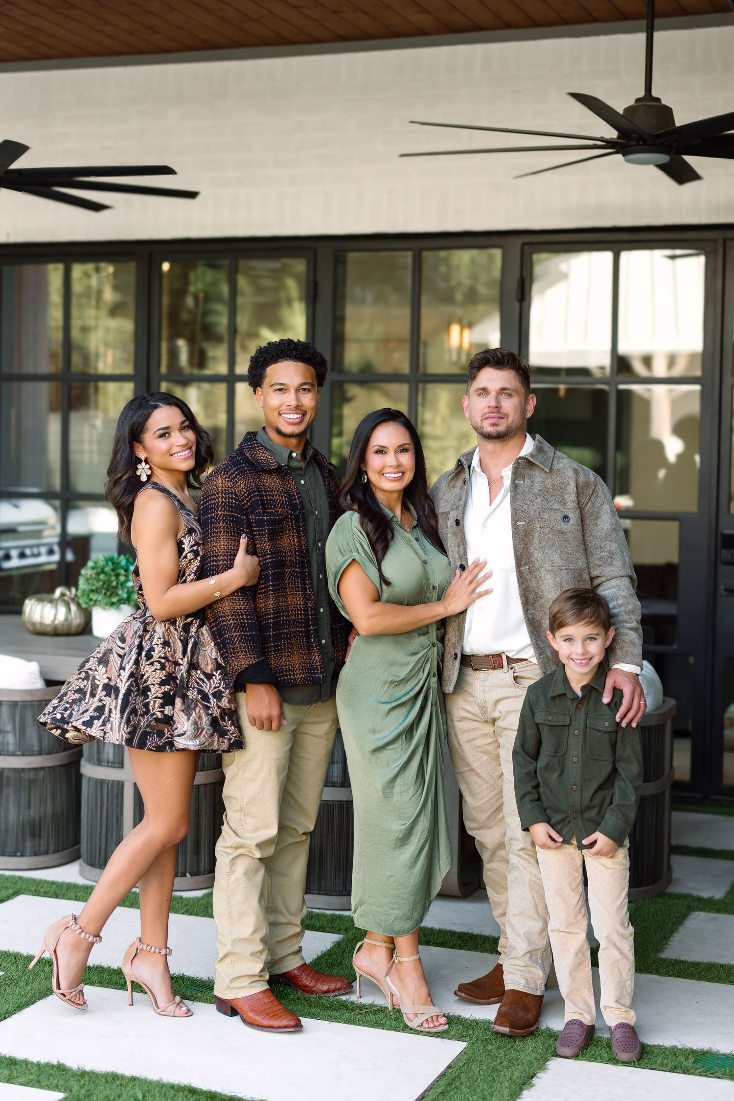 A family of five posing together outdoors in front of a modern home, dressed in coordinated earth-tone outfits.