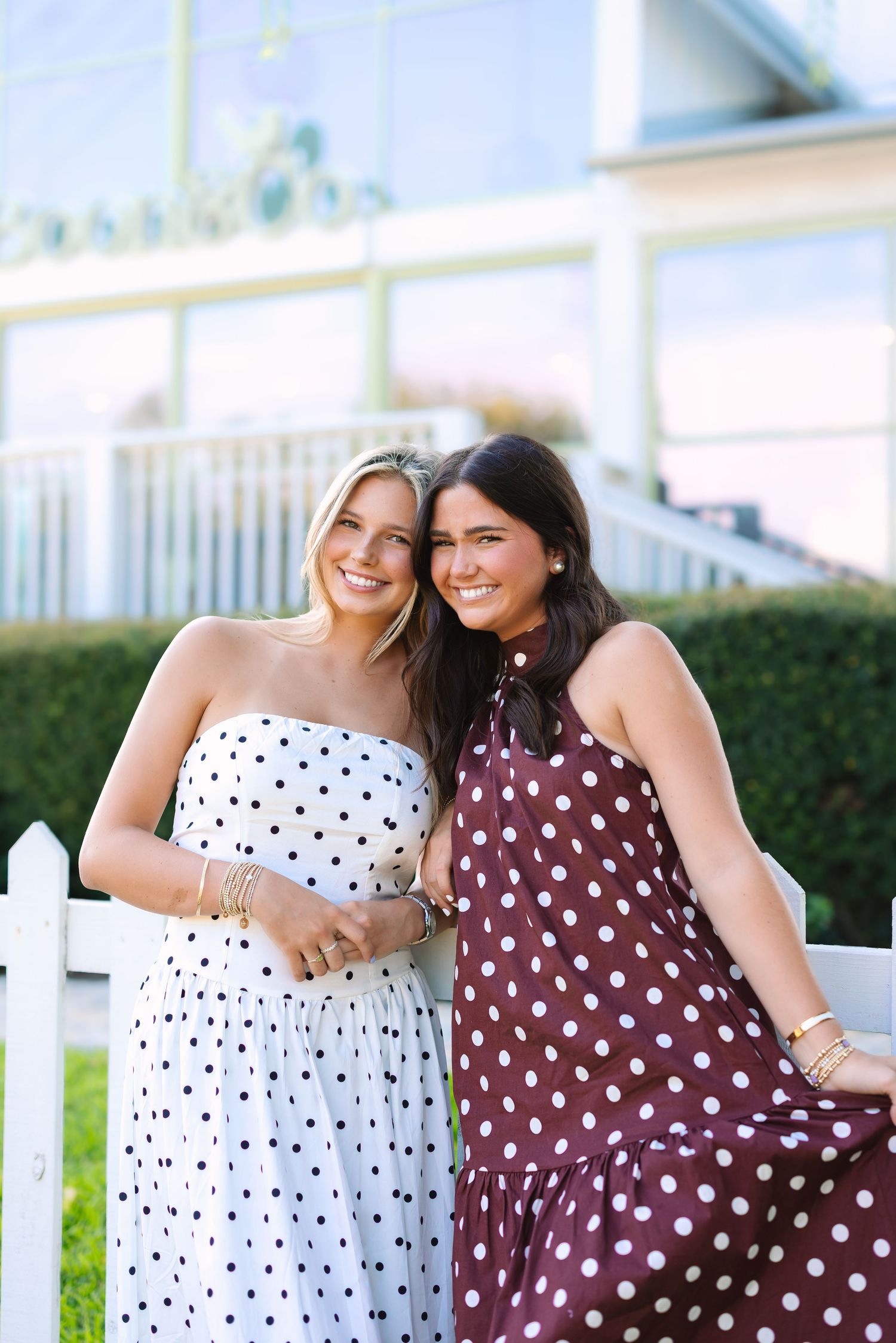 Two friends in polka dot dresses share laughs while leaning on a white fence.