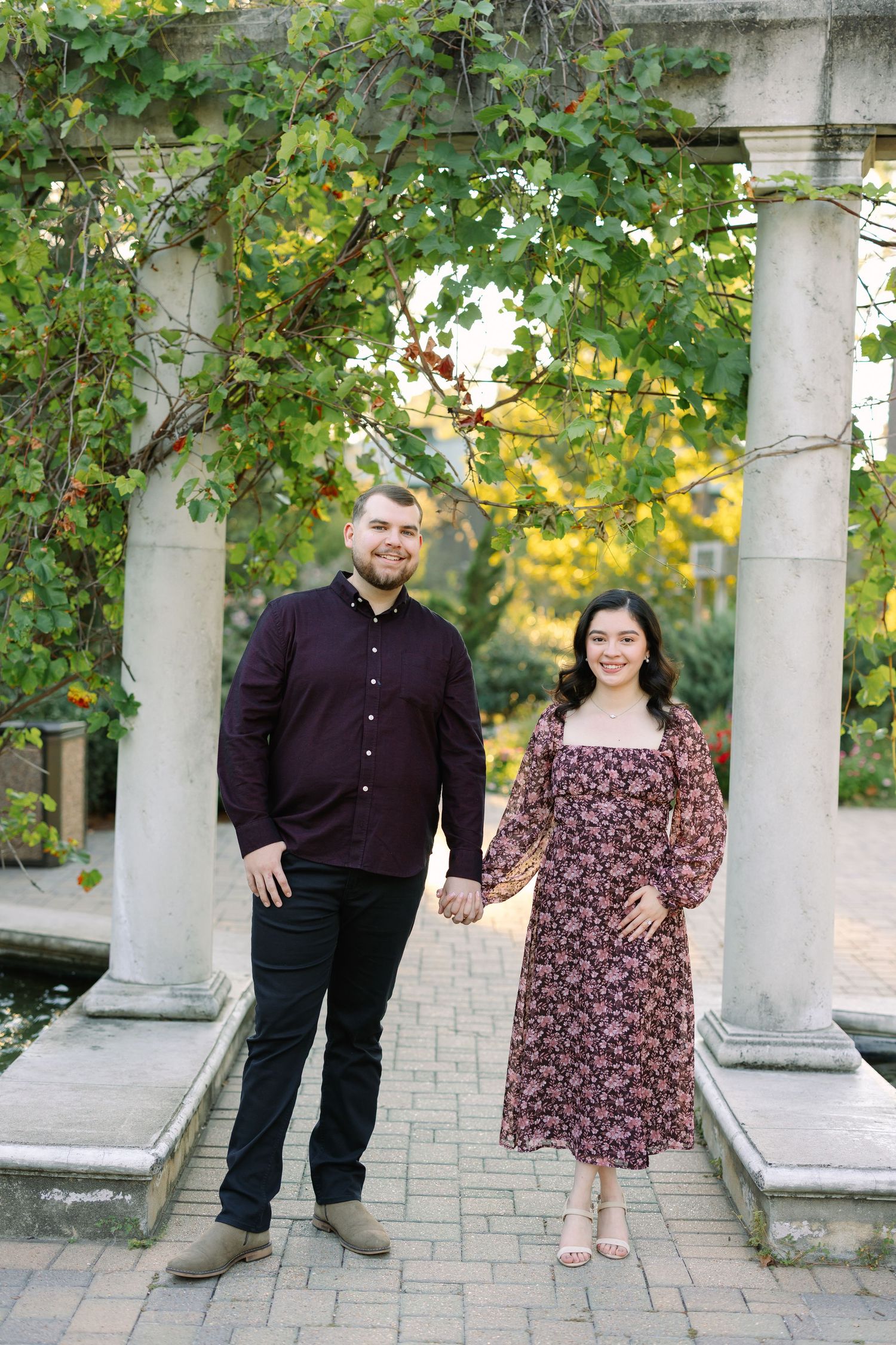 A couple holds hands smiling in a garden with stone columns, he in burgundy shirt, she in floral midi dress.
