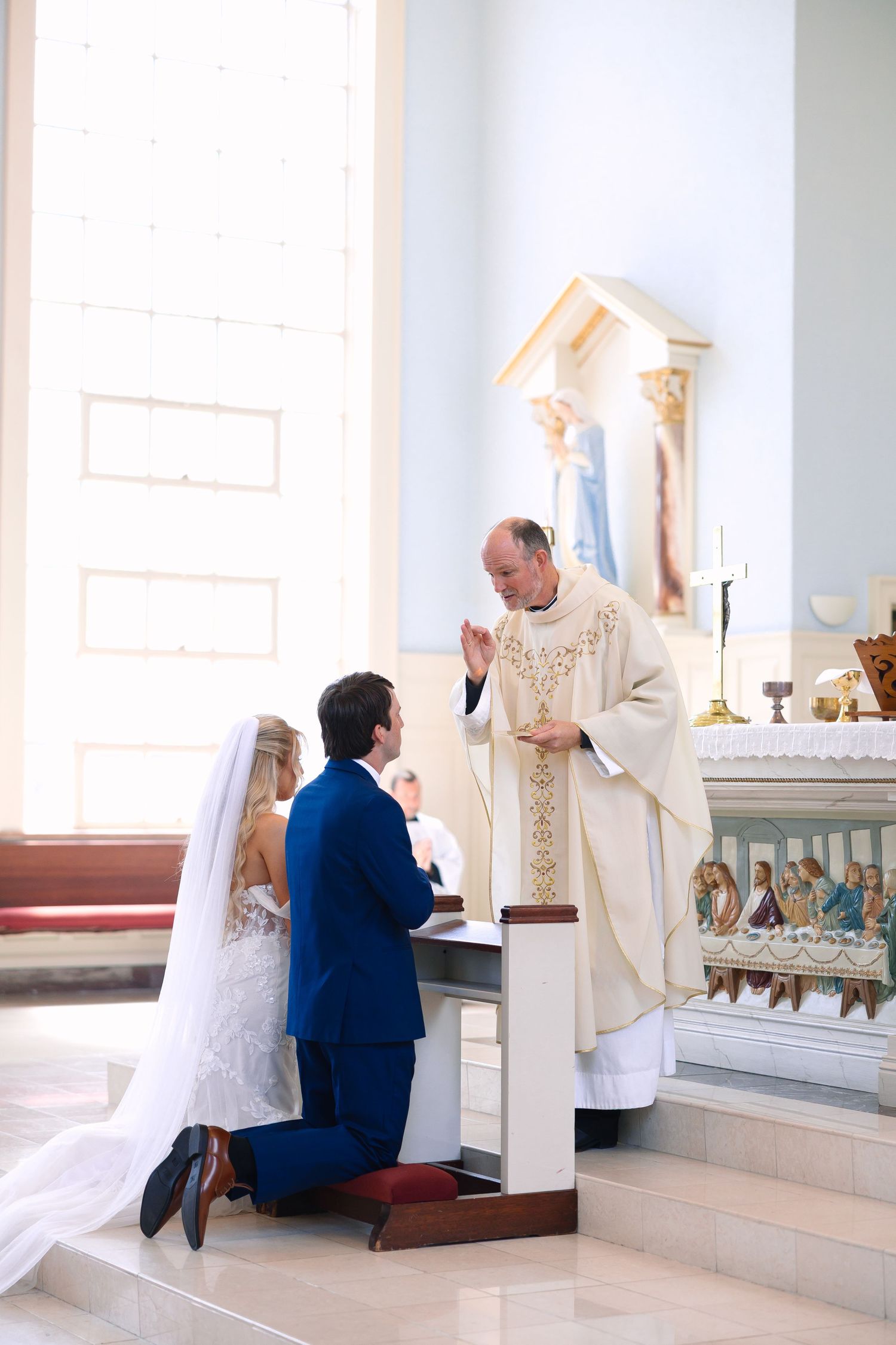 A bride and groom kneel before a priest at the altar during a traditional Catholic wedding ceremony in a bright church.