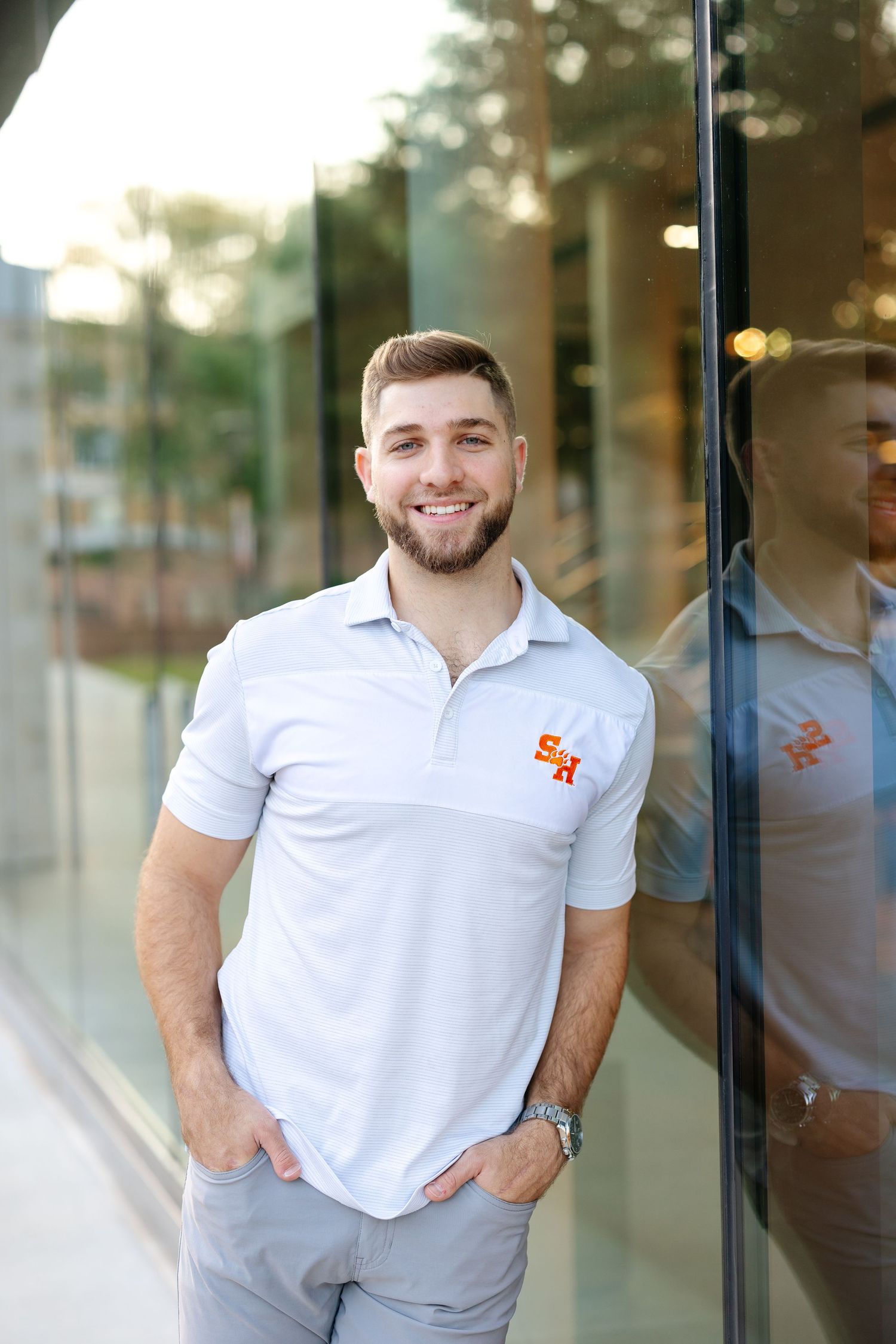 A person in a white polo shirt with a small logo smiles while posing against a reflective glass storefront.