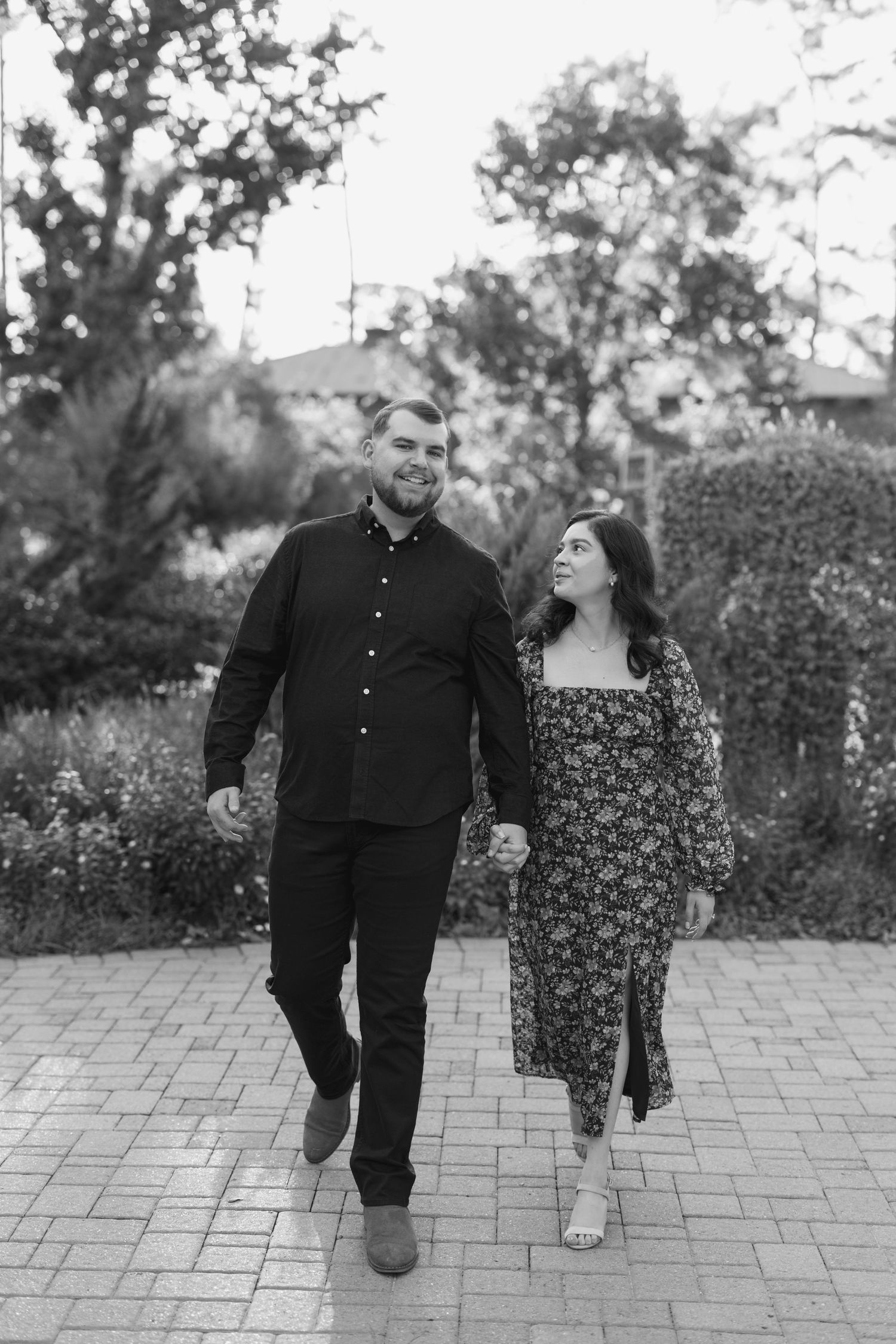 Black and white photo of a couple holding hands and walking together on a brick path in a lush garden setting.