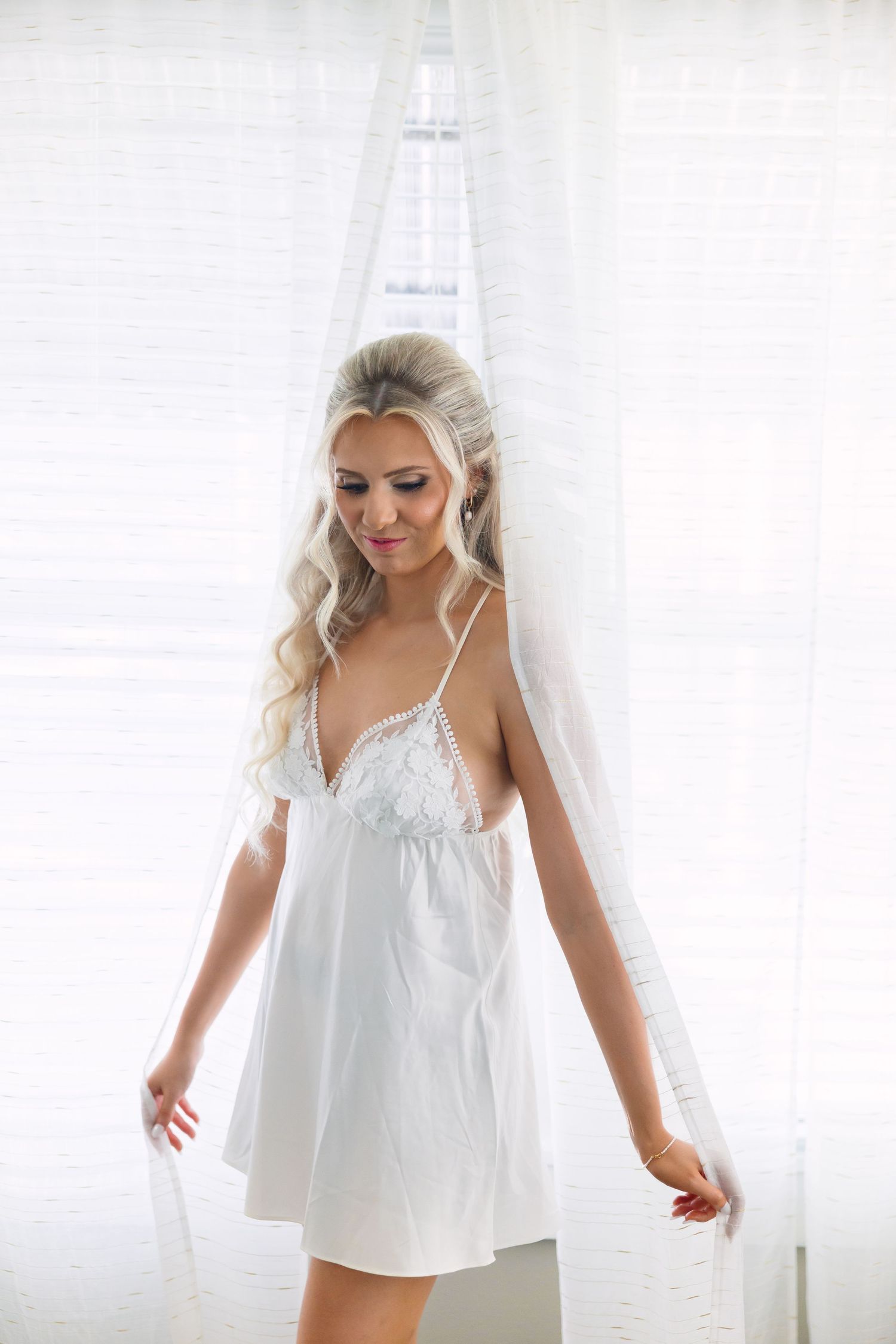 Elegant blonde bride in white lace slip dress stands gracefully by sheer curtained window.