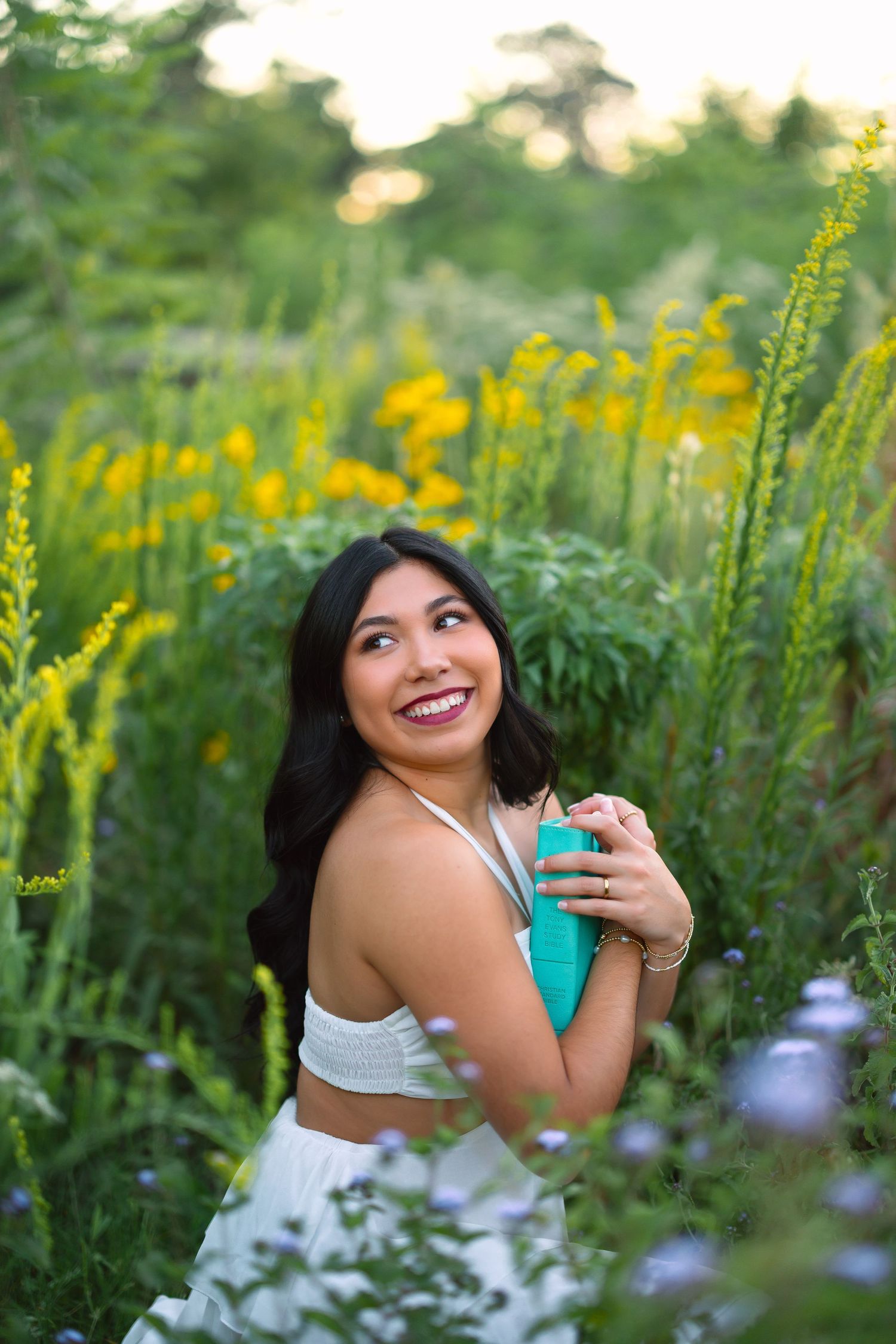 Woman in white outfit smiles while holding turquoise bottle among wildflowers in nature.