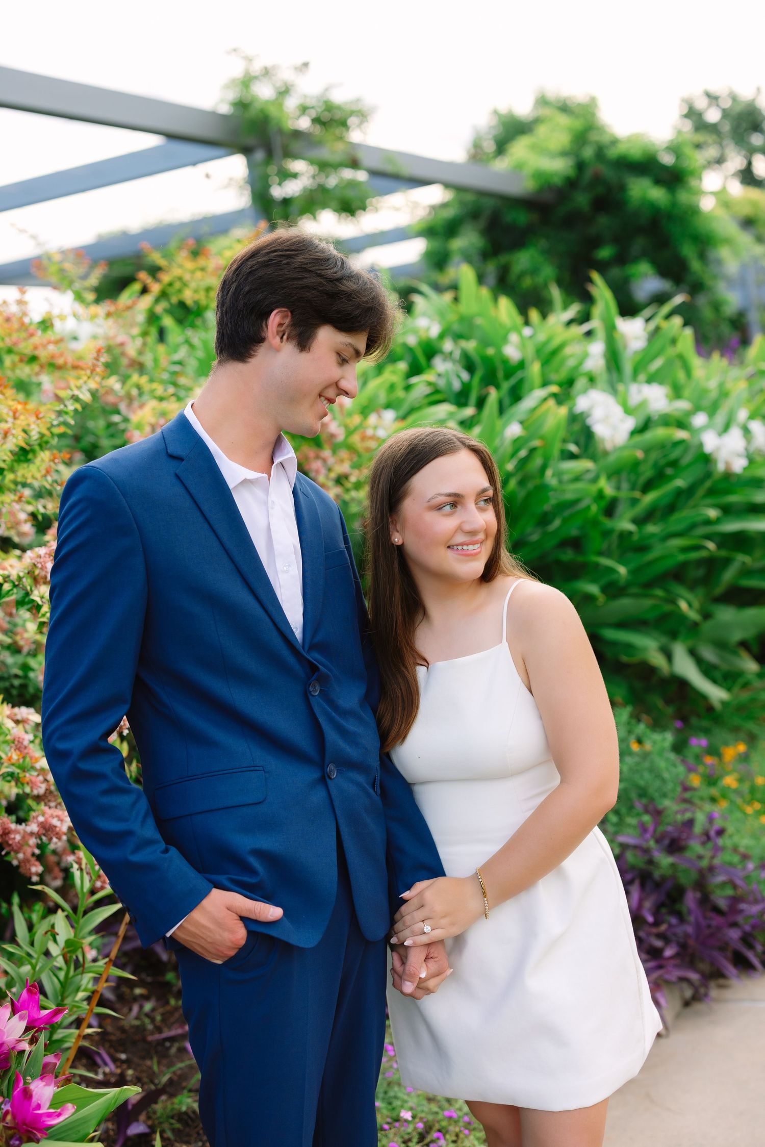 Couple poses together in formal attire among flowering garden plants.